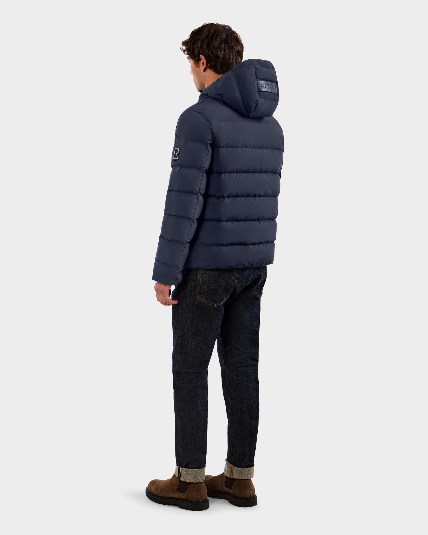 A man faces away from the camera in a Rudsak DEMIAN M - M123507 Navy men’s down puffer, made from sustainable recycled fabric, styled with dark jeans and brown shoes against a plain light gray background.