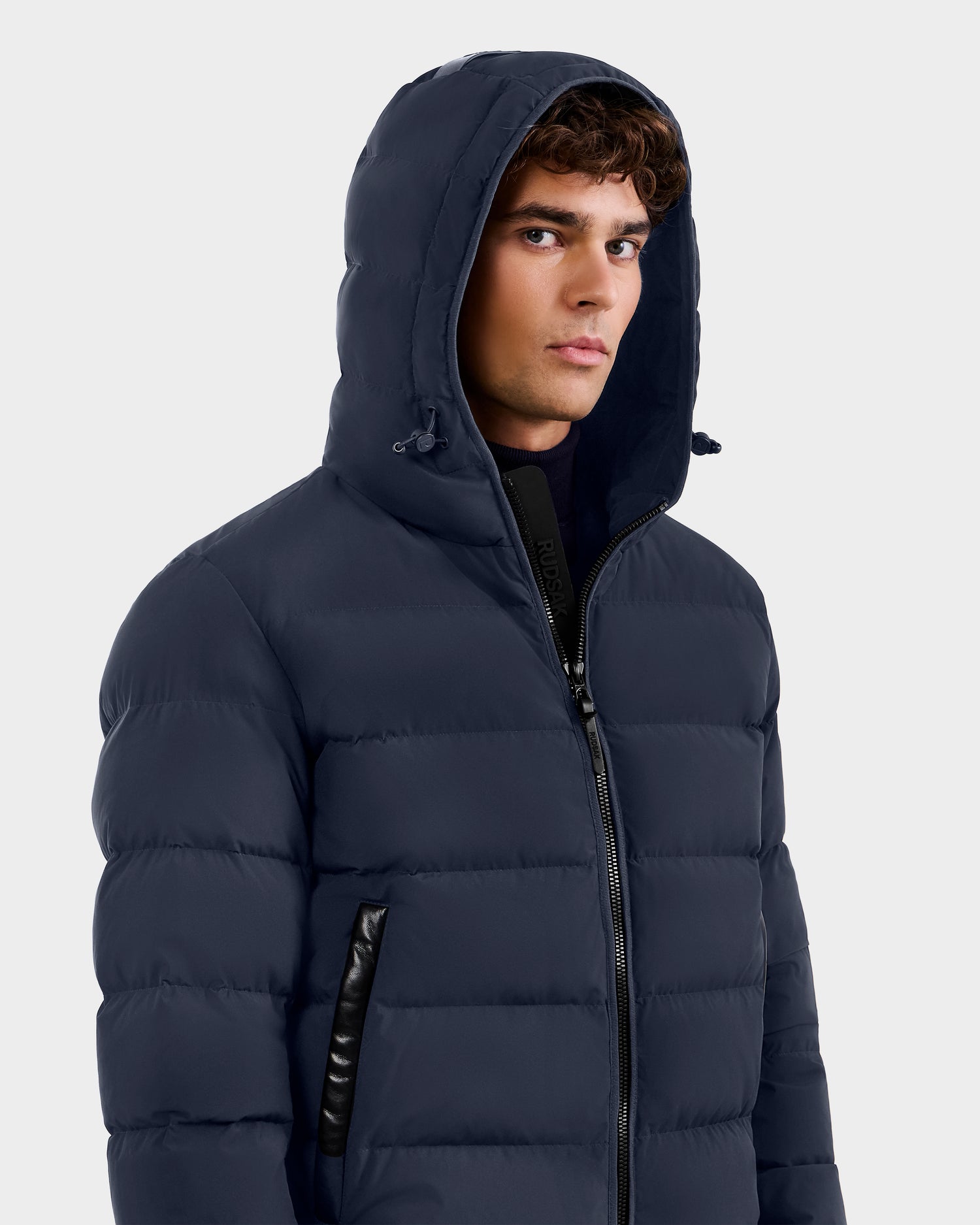 A young man wears the Rudsak DEMIAN M - M123507 Navy down puffer jacket, featuring sustainable recycled fabric, a zipper, and hood, as he stands against a plain light background looking slightly to the side.