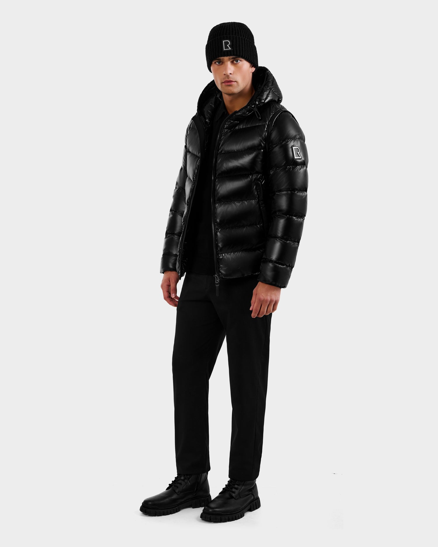A person stands facing forward wearing the Rudsak DEMIAN S-R - M123505 Black RDS-certified down men's puffer jacket, a black beanie, black pants, and black boots against a plain light background.