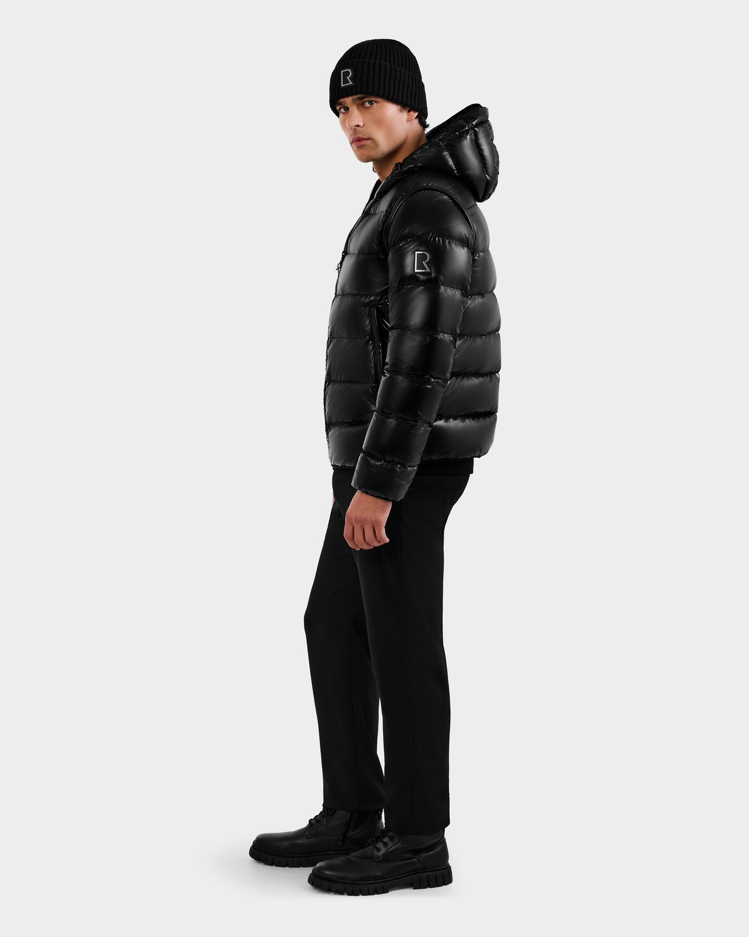 A man wearing the Rudsak DEMIAN S-R - M123505 Black down puffer jacket, along with a black beanie, pants, and boots, stands turned slightly sideways against a plain light gray background.