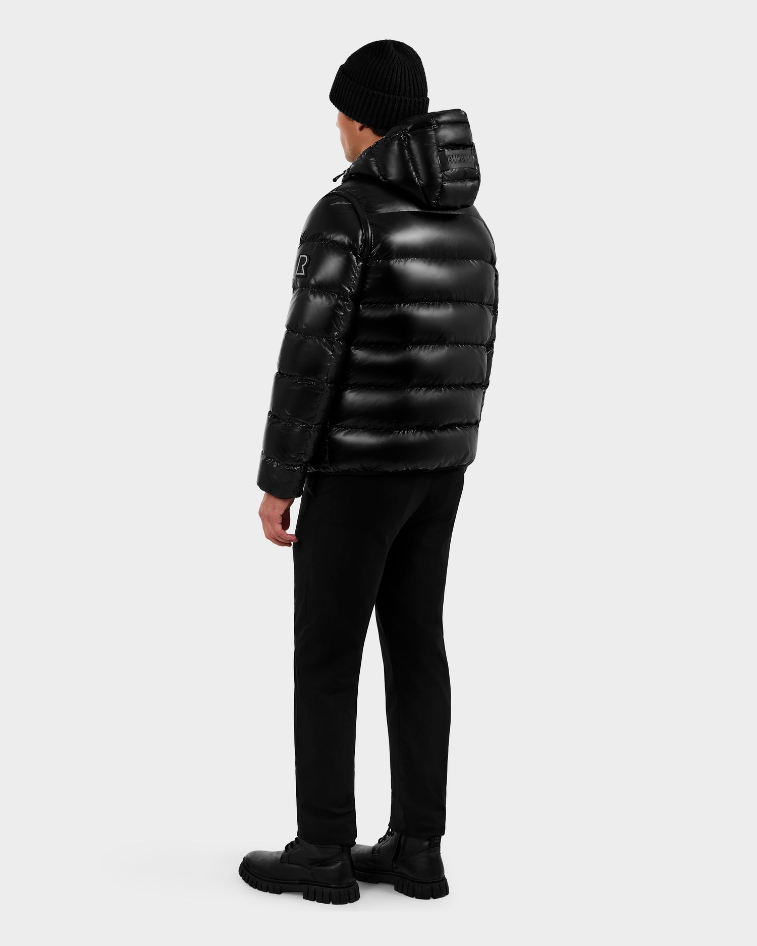 A person faces away against a plain white background, wearing the Rudsak DEMIAN S-R - M123505 Black men's shiny black down puffer jacket with hood, paired with black pants, boots, and a knit beanie.