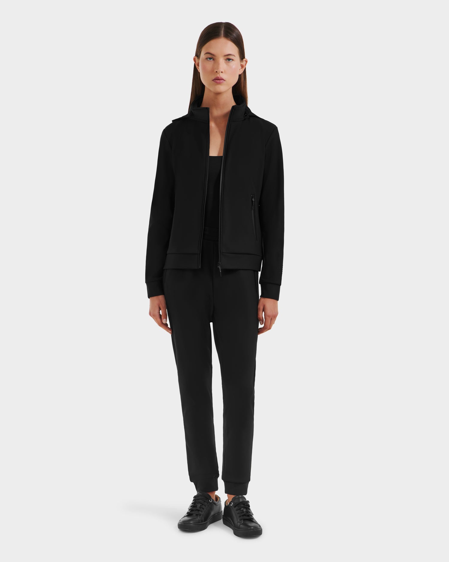 A woman faces forward against a white background, wearing Rudsak’s DERIAM - 8525573 Black sporty jacket with matching stretch pants and black sneakers.