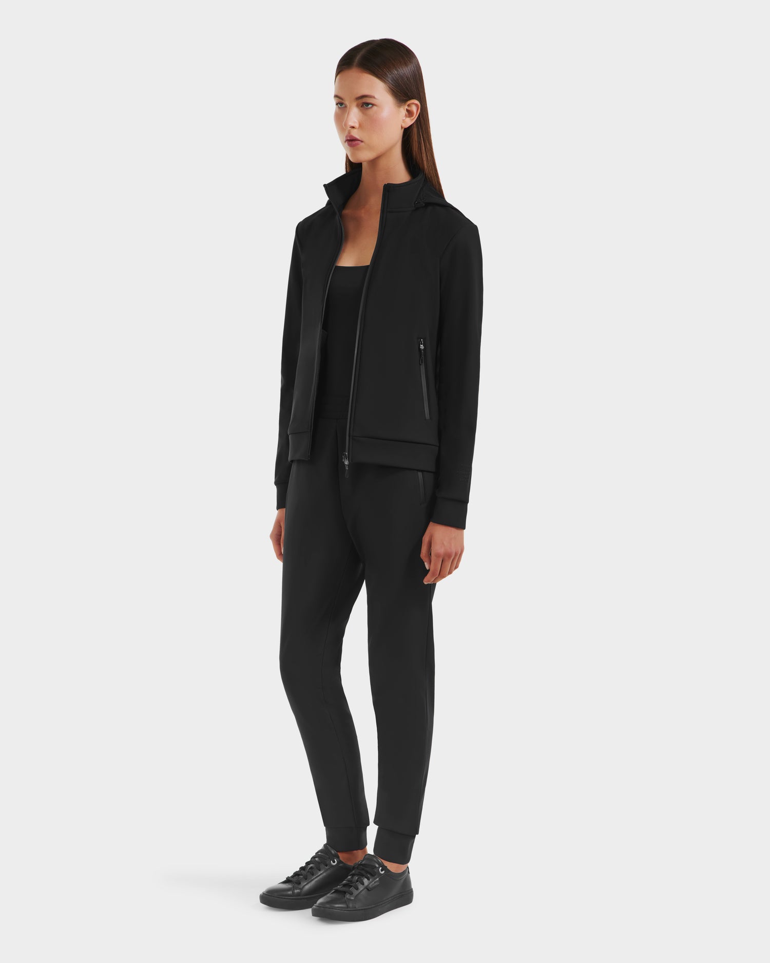 A woman with straight dark brown hair and a neutral expression stands against a white background, wearing the Rudsak DERIAM - 8525573 Black women’s zip-up sweater with performance stretch fabric, black top, pants, and sneakers.