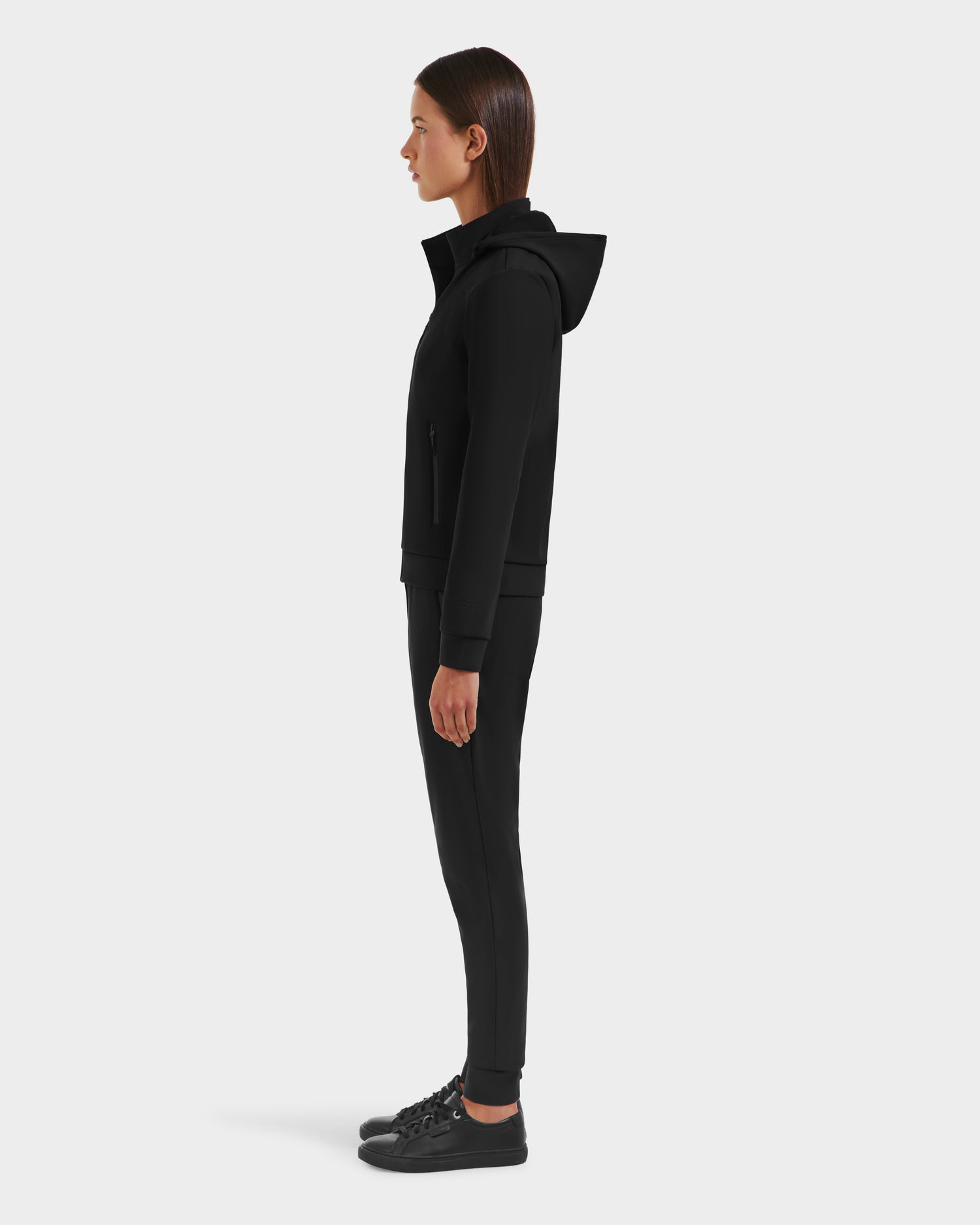 A person stands in profile facing left, wearing the Rudsak DERIAM - 8525573 Black jacket with black pants and sneakers, against a light gray background.