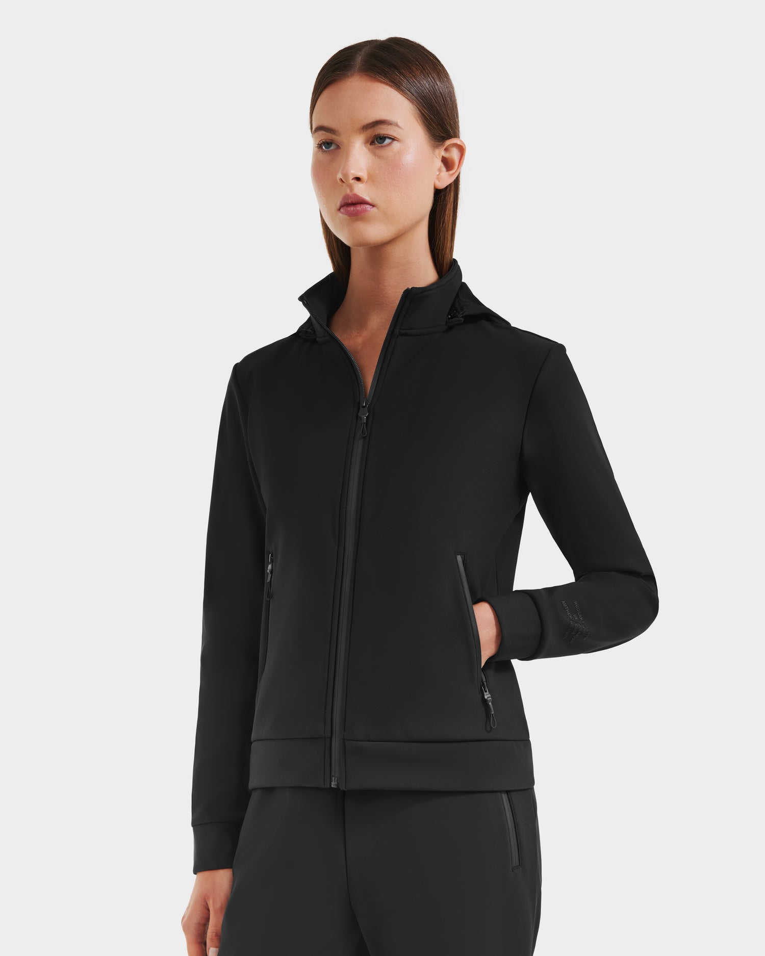 A woman with straight brown hair and a neutral expression stands against a plain background, wearing the Rudsak DERIAM - 8525573 Black sporty zip-up sweater jacket and matching pants, one hand in her pocket.