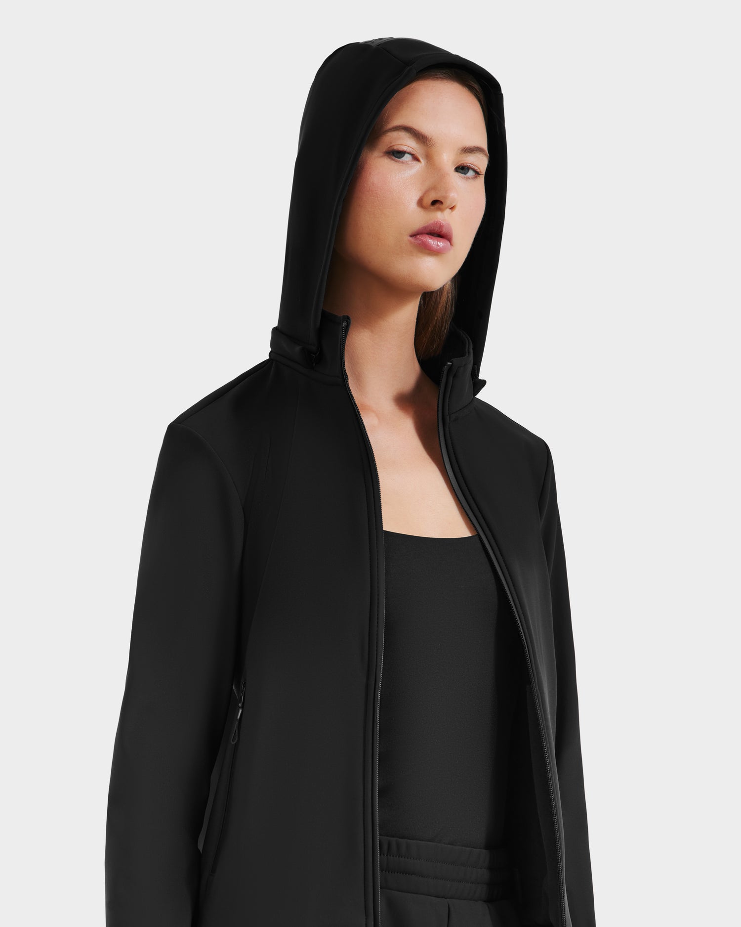 A woman wears the Rudsak DERIAM - 8525573 Black jacket with performance stretch fabric, hood up and unzipped over a black top, confidently facing the camera against a plain light background.