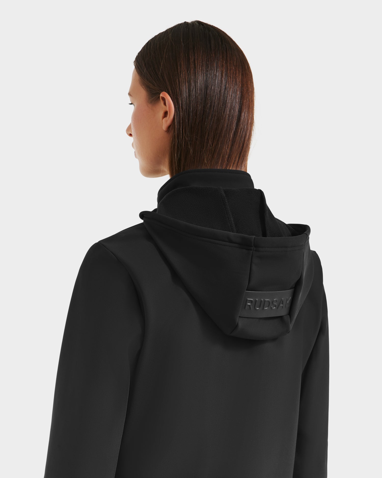 A person with straight brown hair is shown from the back wearing the Rudsak DERIAM - 8525573 Black sporty hooded jacket, featuring performance stretch fabric and a raised RUDSAK logo on the hood, against a plain light background.