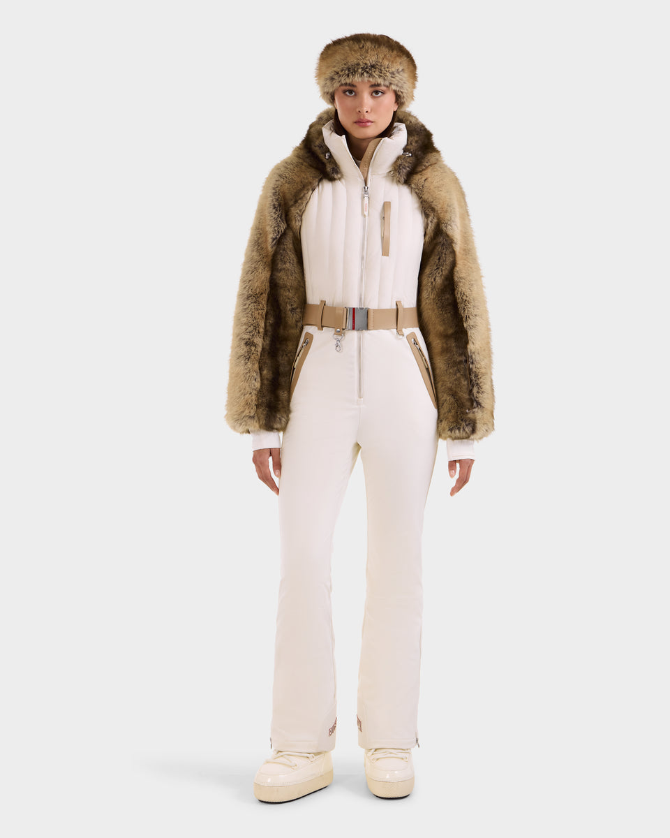 A woman faces forward in a Rudsak DESTINY - 8124727 Vanilla women’s ski suit with tan belt, faux fur jacket draped over her shoulders, matching faux fur hat, and white platform boots against a plain background.