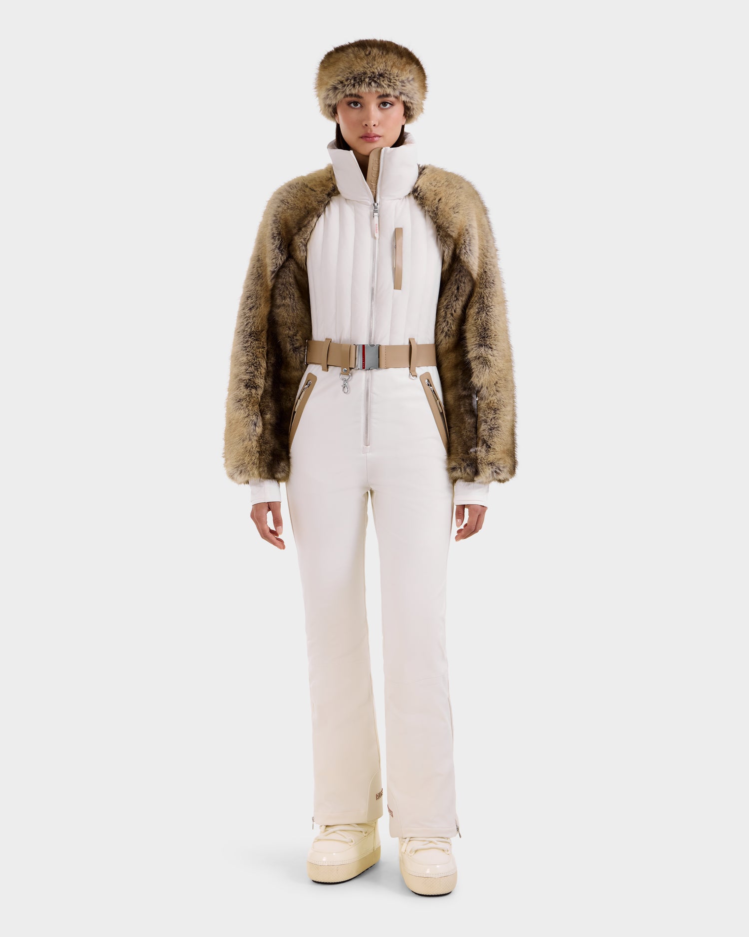 A woman stands facing forward in the Rudsak DESTINY - 8124727 Vanilla, a white one-piece ski suit with fur-trimmed jacket, matching fur hat, beige belt, and white boots, set against a plain white background.