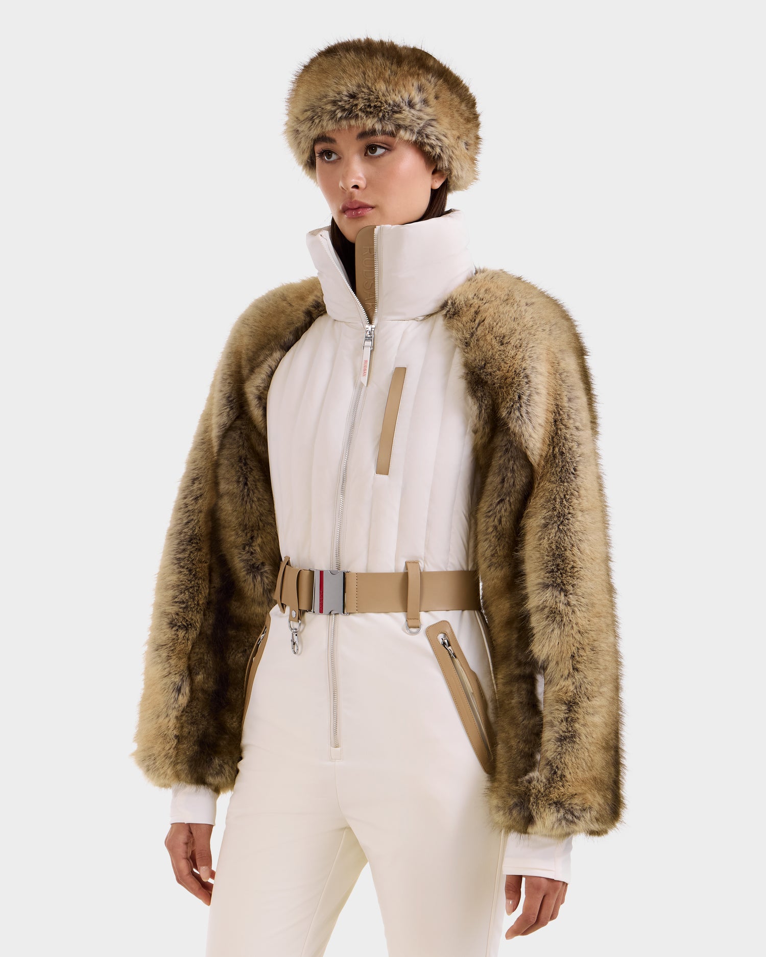 A woman in the Rudsak DESTINY - 8124727 Vanilla white one-piece ski suit with a belted waist and high collar, styled with a brown faux fur jacket and matching hat, stands against a plain light background.