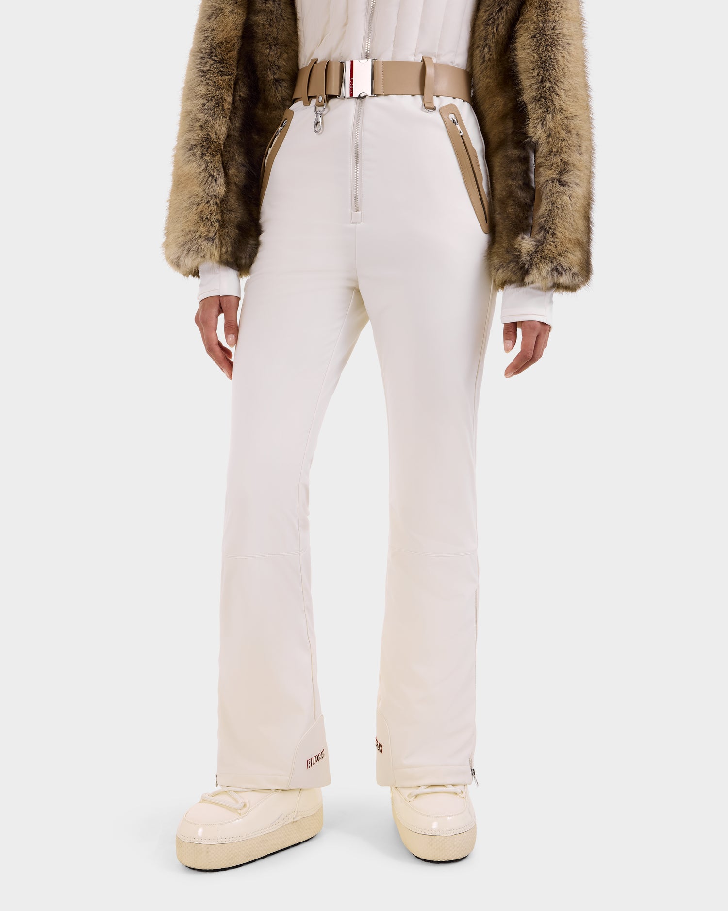 A person wears the Rudsak DESTINY - 8124727 Vanilla women's ski suit with flared pants, zippered pockets, a tan belt, beige fur sleeves, chunky white boots, and RECCO rescue technology, standing against a plain white background.