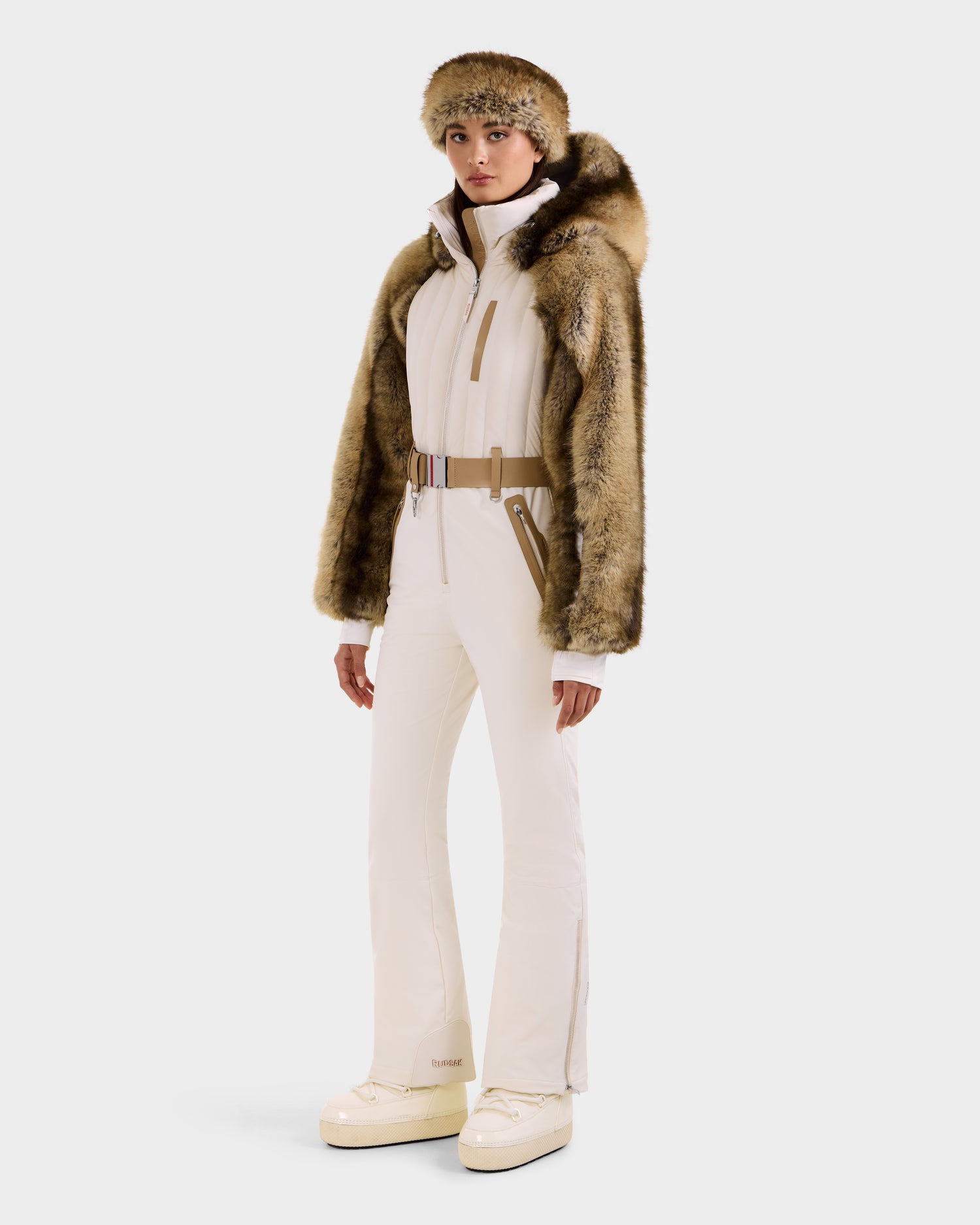 A person stands against a plain light background wearing the Rudsak DESTINY - 8124727 Vanilla women's one-piece ski suit with brown accents, a matching faux fur jacket and hat, and white platform boots.