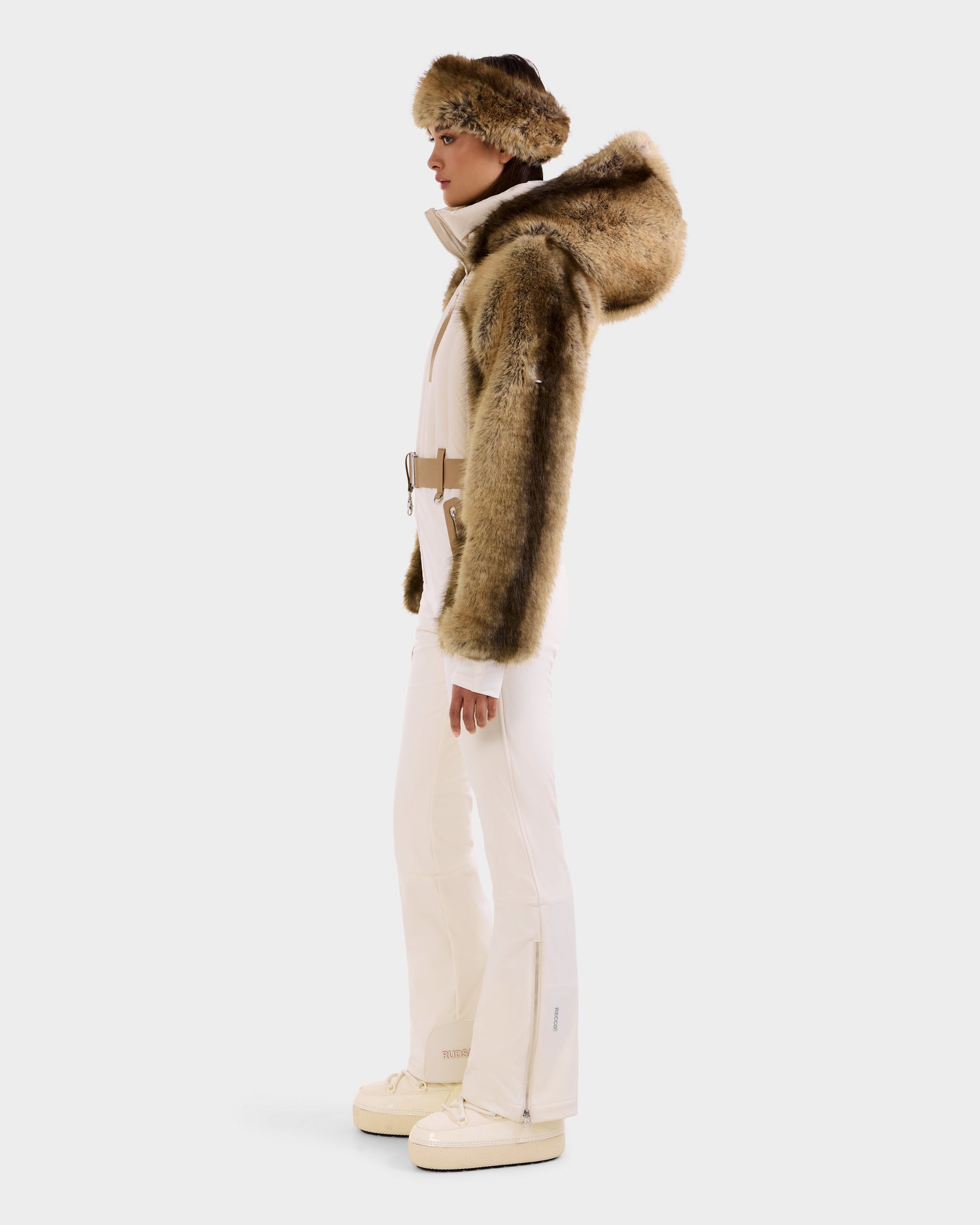 Wearing the Rudsak DESTINY - 8124727 Vanilla one-piece ski suit with flared pants, beige belt, faux fur-trimmed hood, and cream boots, a person in profile stands out stylishly against a plain white background.
