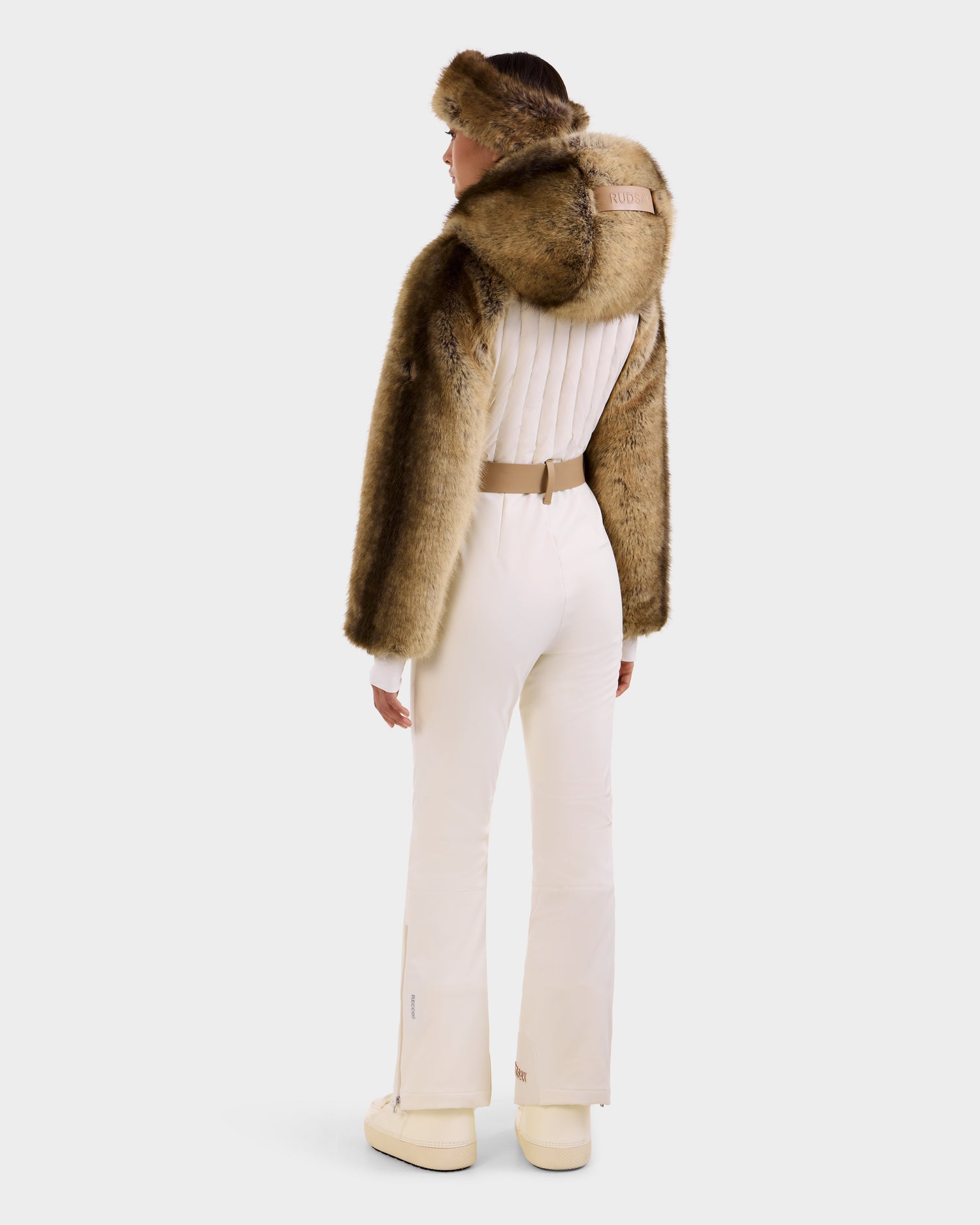 A woman stands with her back to the camera, wearing the Rudsak DESTINY - 8124727 Vanilla fitted women's ski suit with a tan fur cape and matching headband, paired with white boots against a light background.