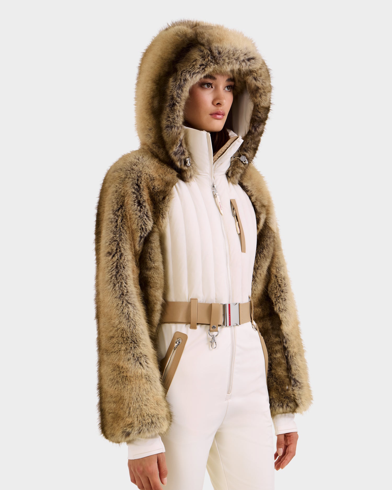 A person wearing the Rudsak DESTINY - 8124727 Vanilla, a tan and brown faux fur hooded jacket, over a white padded women's ski suit with belted waist and tan zipper accents, stands against a plain light background.