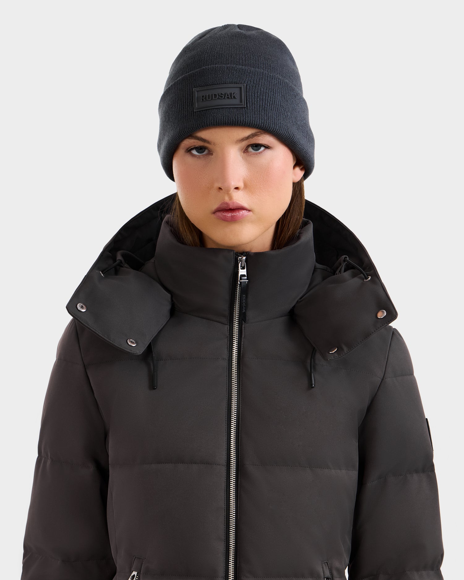 A young woman in a dark Rudsak DILLION - U423501 Grey puffer jacket with a high collar and a unisex knit hat looks directly at the camera with a neutral expression against a plain light background.