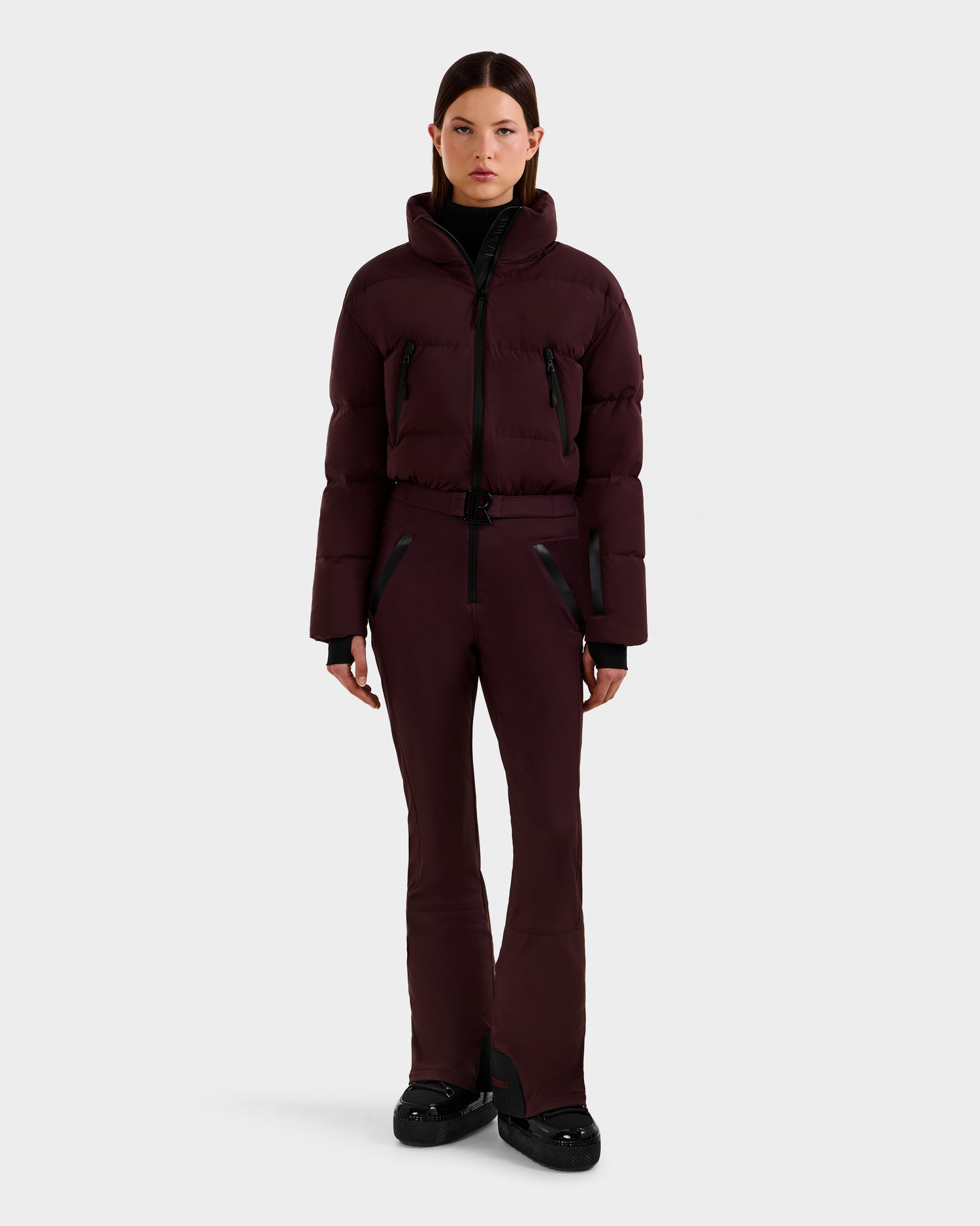 A person stands facing forward in the Rudsak DINARA - 8125600 Bordeaux women’s ski suit with black accents and black shoes, featuring RECCO rescue technology, against a plain white background.