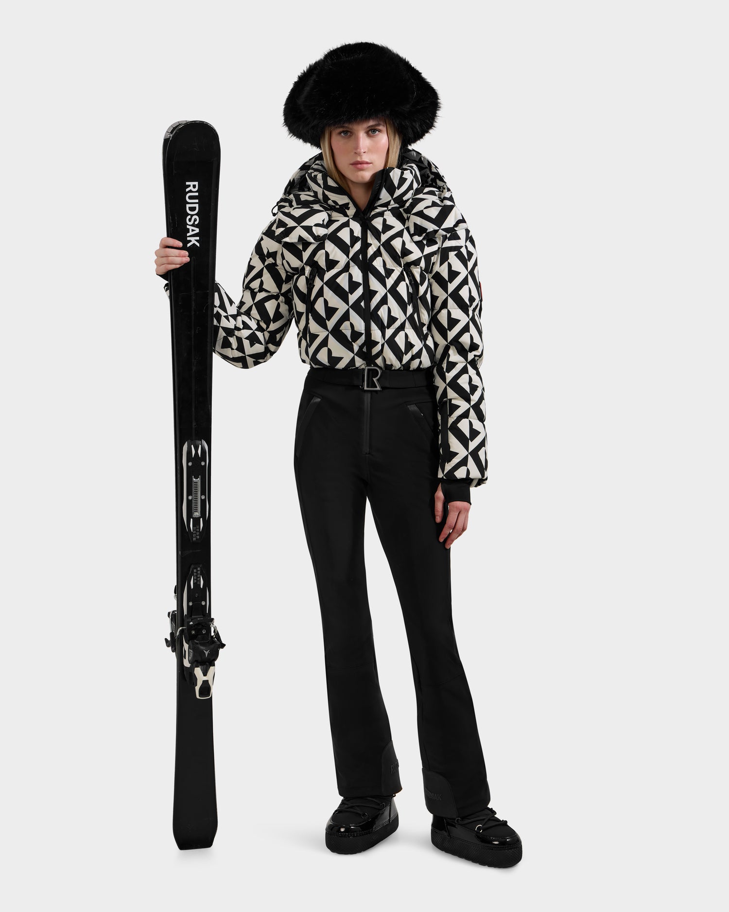 A person stands holding black skis vertically, wearing the Rudsak DINARA - 8125600 Vanilla Monogram women’s ski suit with black pants, black shoes, and a large black fur hat against a plain white background.
