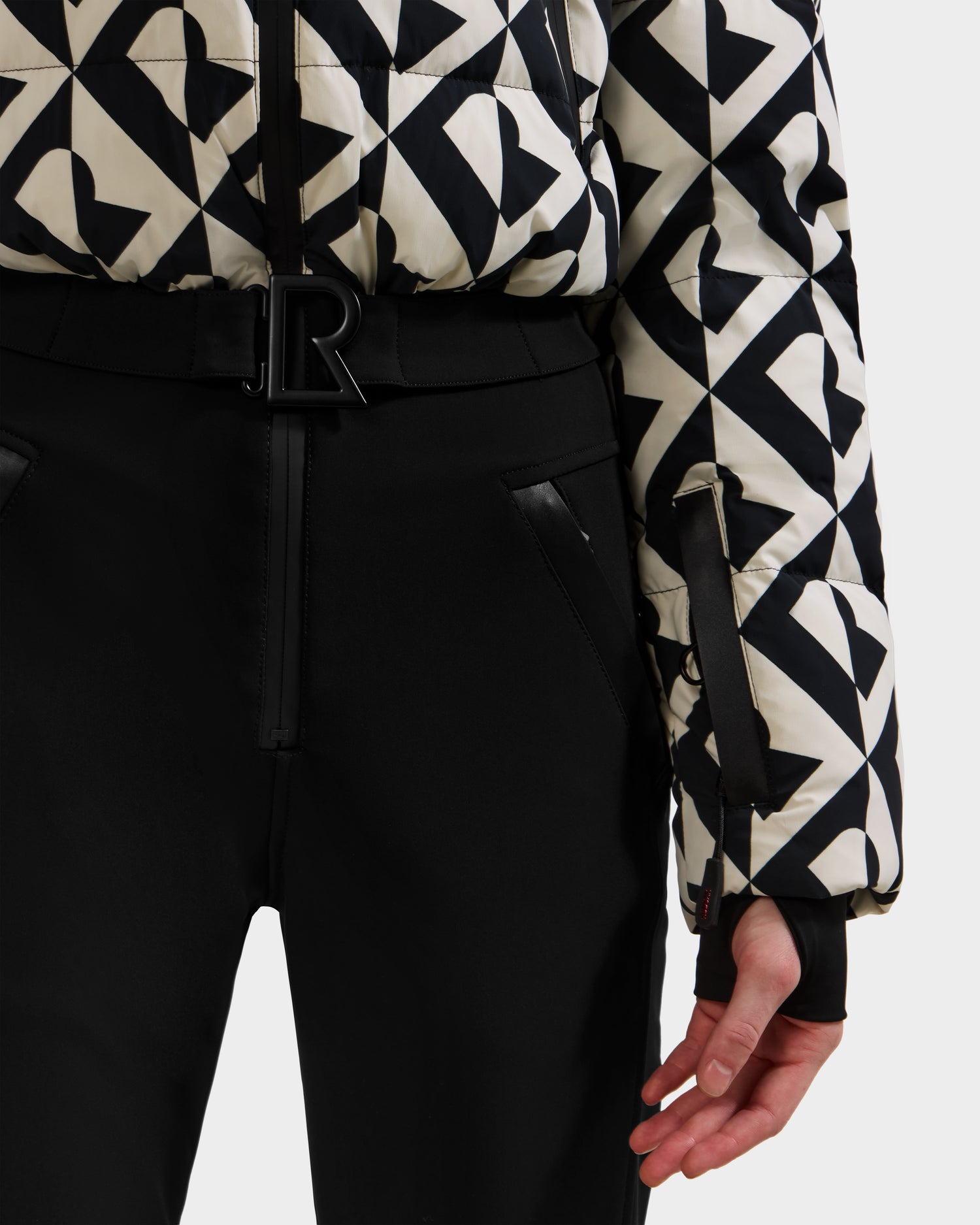 A person wearing Rudsak’s DINARA - 8125600 Vanilla Monogram women’s ski suit with black high-waisted pants and a black belt featuring an R buckle, their left hand relaxed at their side.