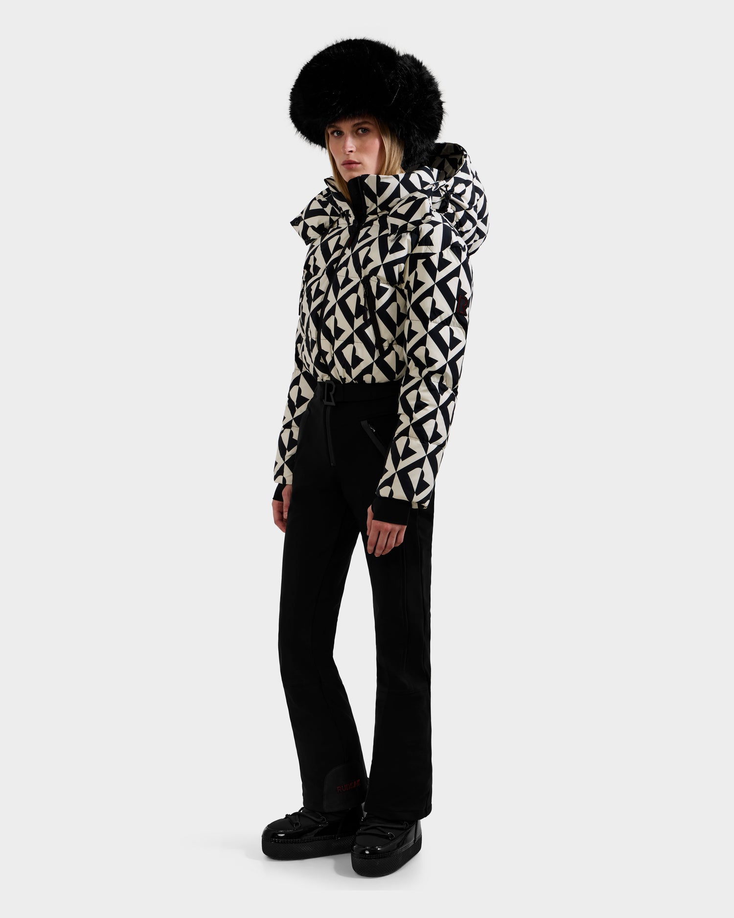 A person stands against a plain background wearing the Rudsak DINARA - 8125600 Vanilla Monogram women’s ski suit, featuring a monogrammed winter jacket, black ski pants, black boots, and a large black fur hat.