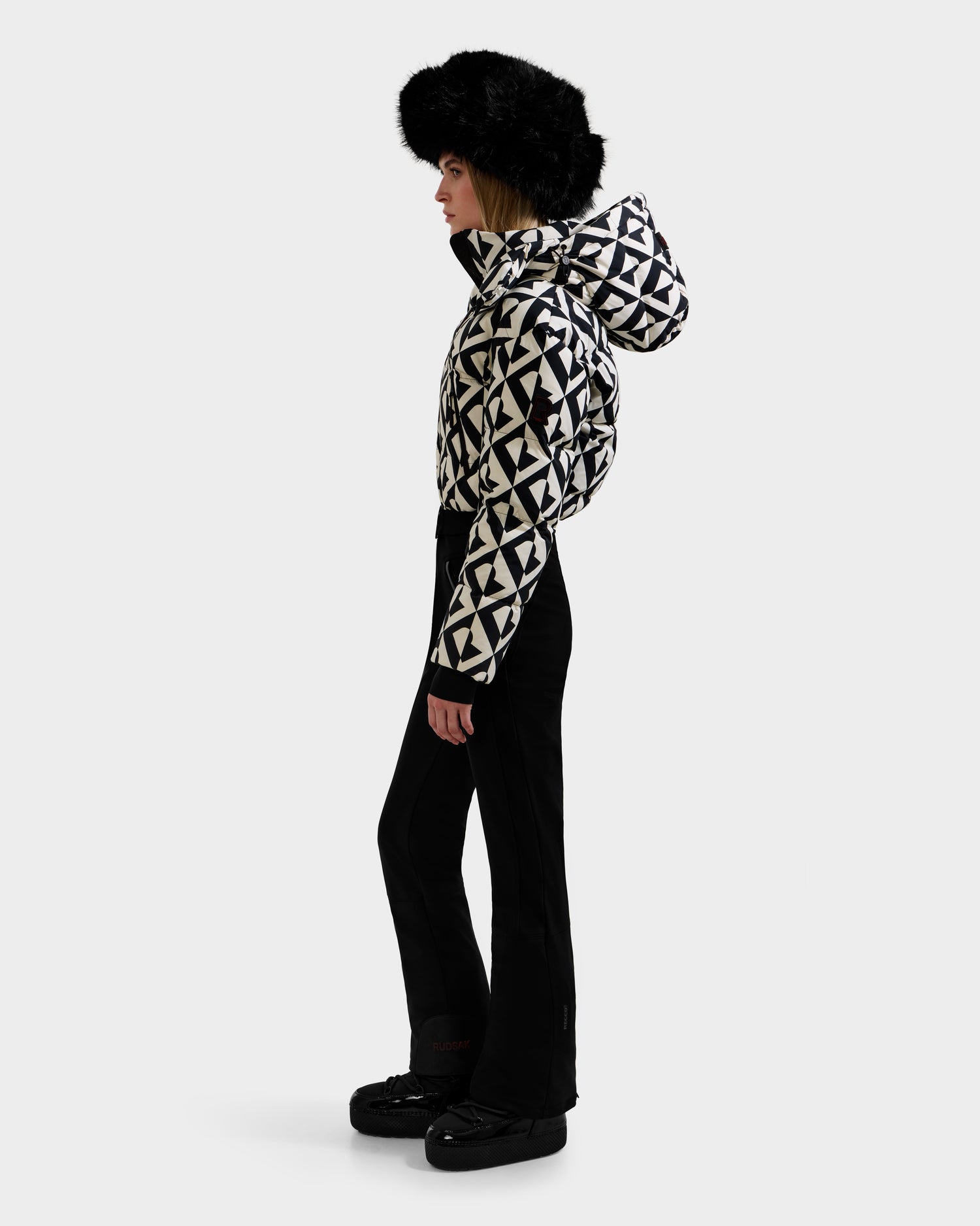 A person stands in profile against a white background wearing Rudsak's DINARA - 8125600 Vanilla Monogram women's ski suit with a hood, paired with black flared pants, black boots, and a large black fur hat.