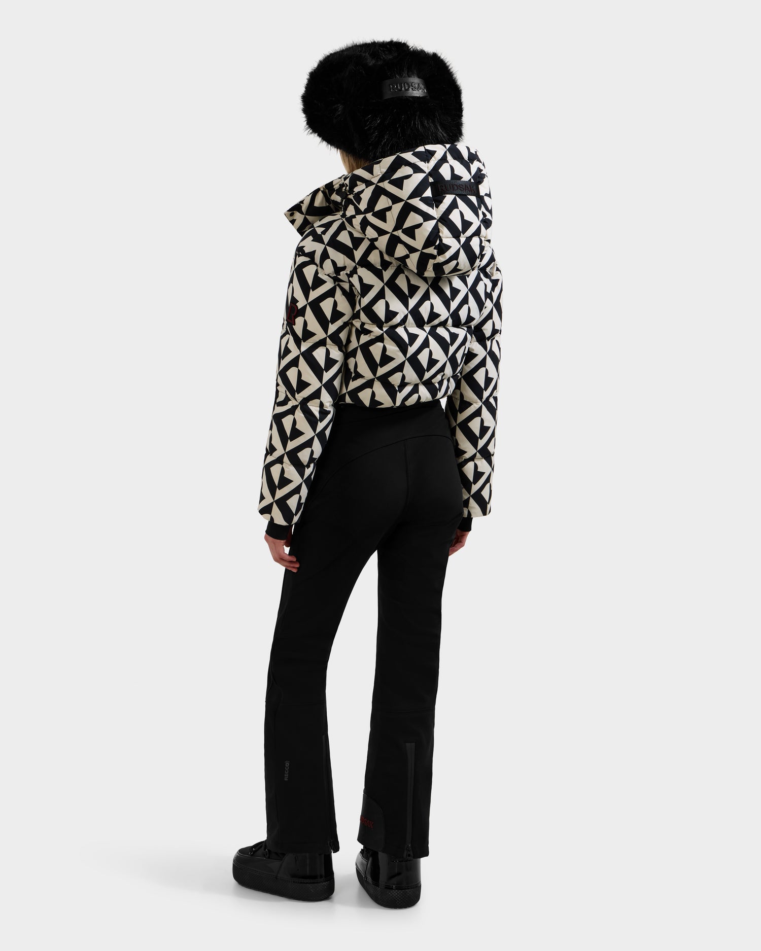 A person faces away, wearing black pants and shoes, and a women's Rudsak DINARA - 8125600 Vanilla Monogram ski suit with a white and black geometric-patterned jacket with black fur-trimmed hood against a plain light background.