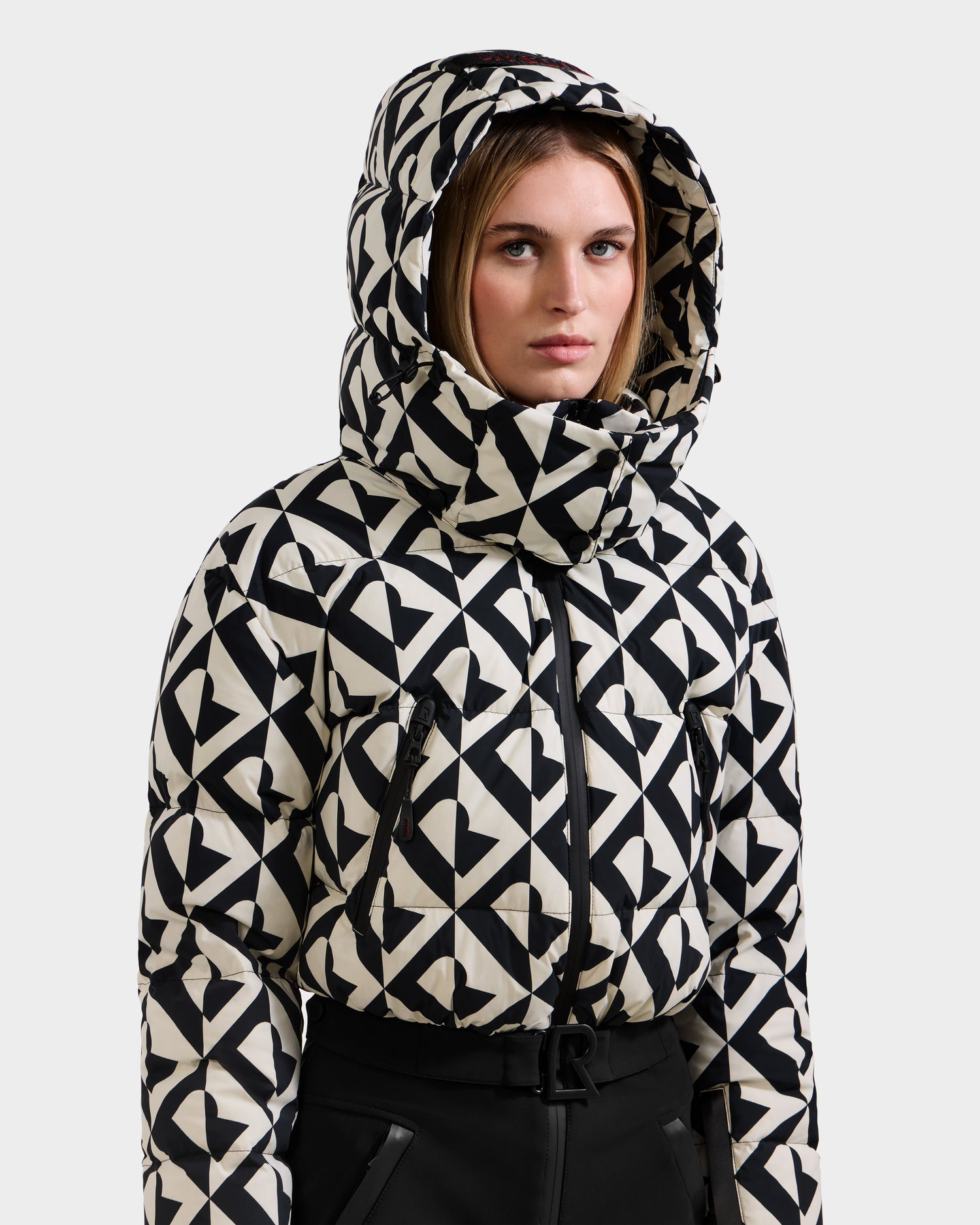A woman stands against a plain light background wearing the Rudsak DINARA - 8125600 Vanilla Monogram hooded puffer jacket with a bold geometric heart pattern and RECCO rescue technology.