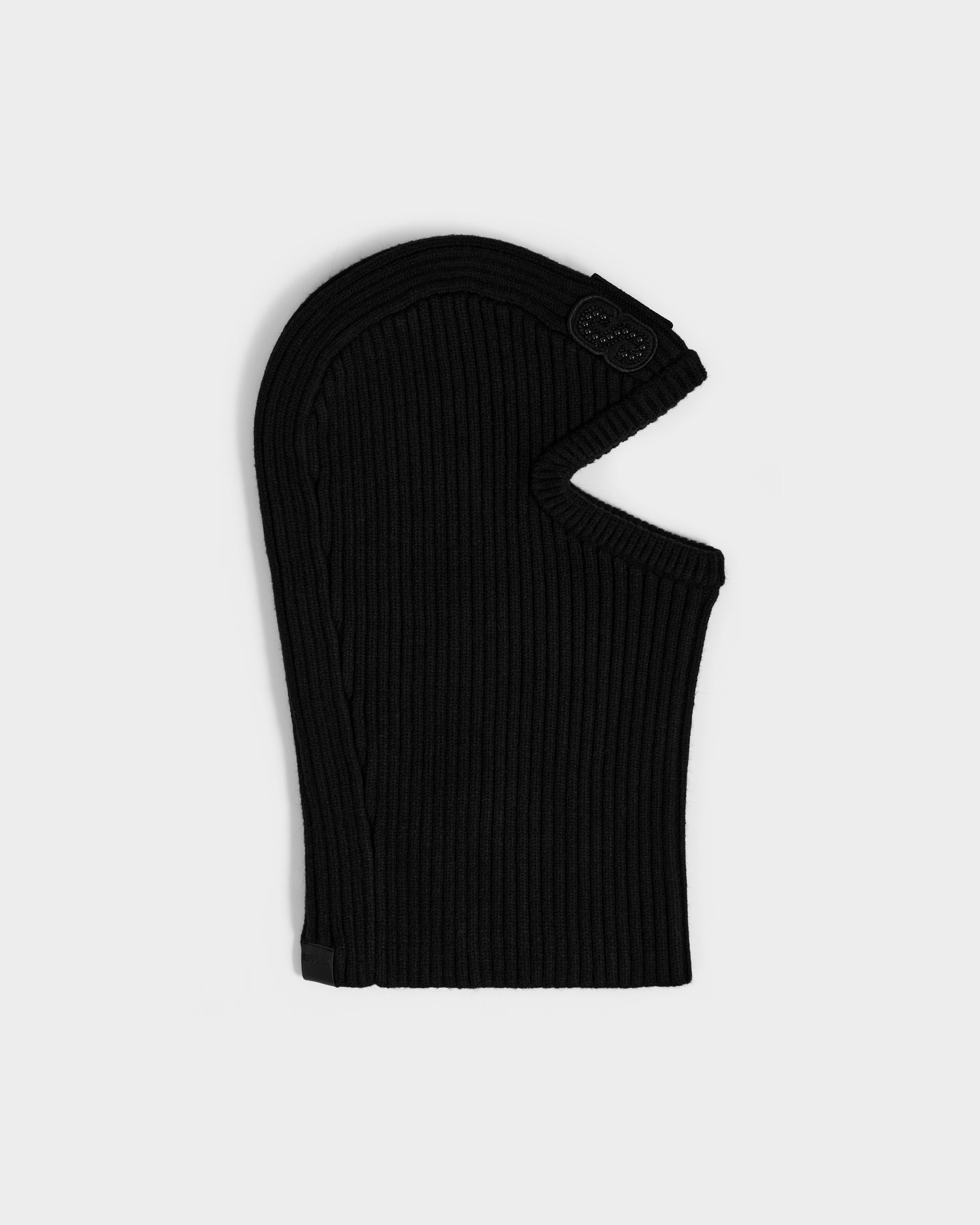 The DIXIE - 8425551 Black by Rudsak is a black, ribbed women’s knit balaclava with an eye and nose opening, ideal for winter wear, shown laid flat on a light gray background.