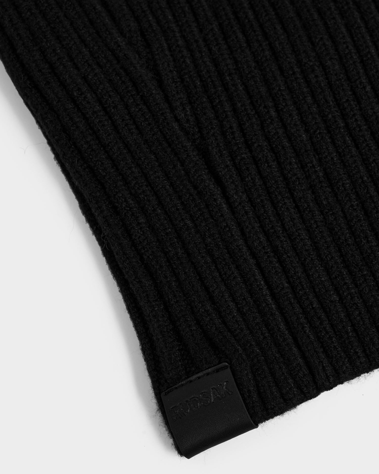Close-up of the DIXIE - 8425551 Black women's knit balaclava by Rudsak, featuring black ribbed fabric and a small black leather tag embossed with RUSLAN, set against a plain white background for a modern winter look.