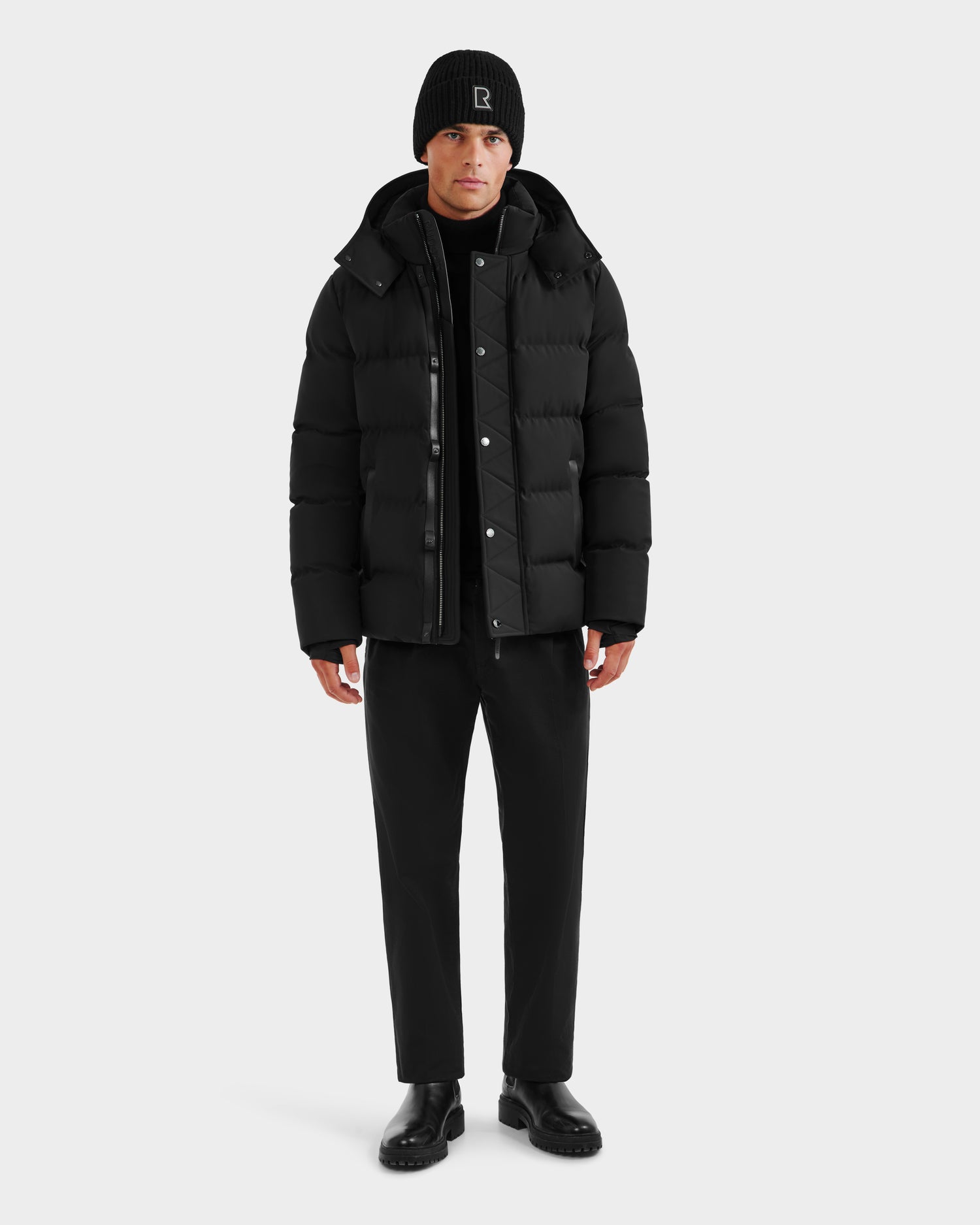 A man faces forward in a Rudsak DOMINIC - 6125041 Black winter jacket with a recycled shell, paired with black pants, boots, beanie, and shirt, against a plain light gray background.