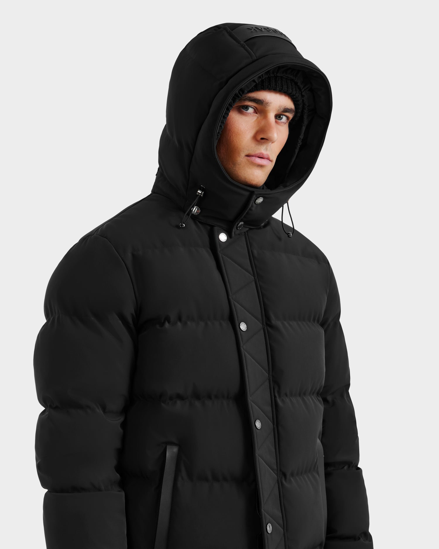A person wearing the Rudsak DOMINIC - 6125041 Black men's down parka with a hood and a black knit beanie, looking slightly to the side against a plain light background.