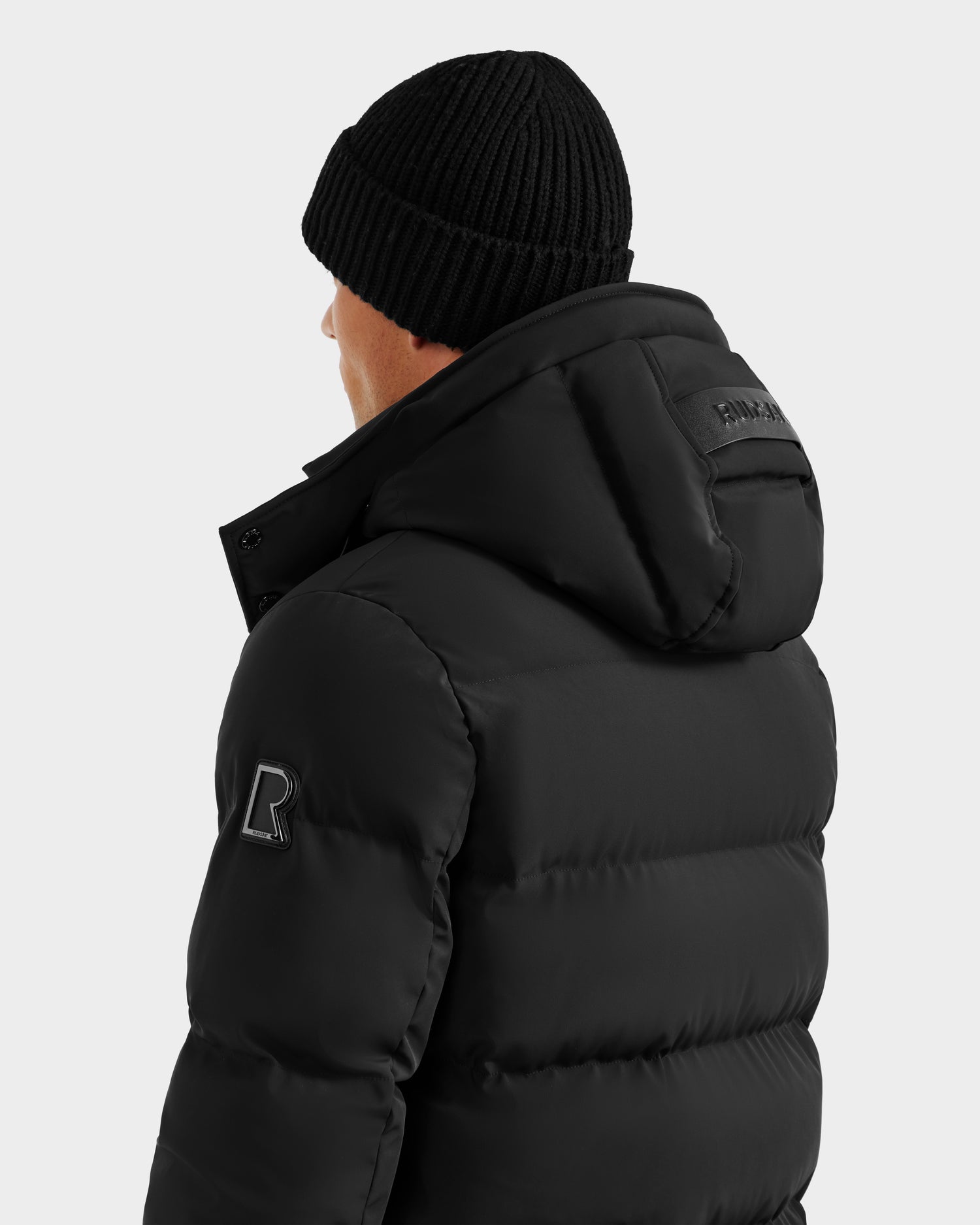 A person is shown from the back and slightly to the side against a plain light background, wearing the Rudsak DOMINIC - 6125041 Black men’s down parka with a recycled shell, visible stitching, sleeve patch, and a black beanie.