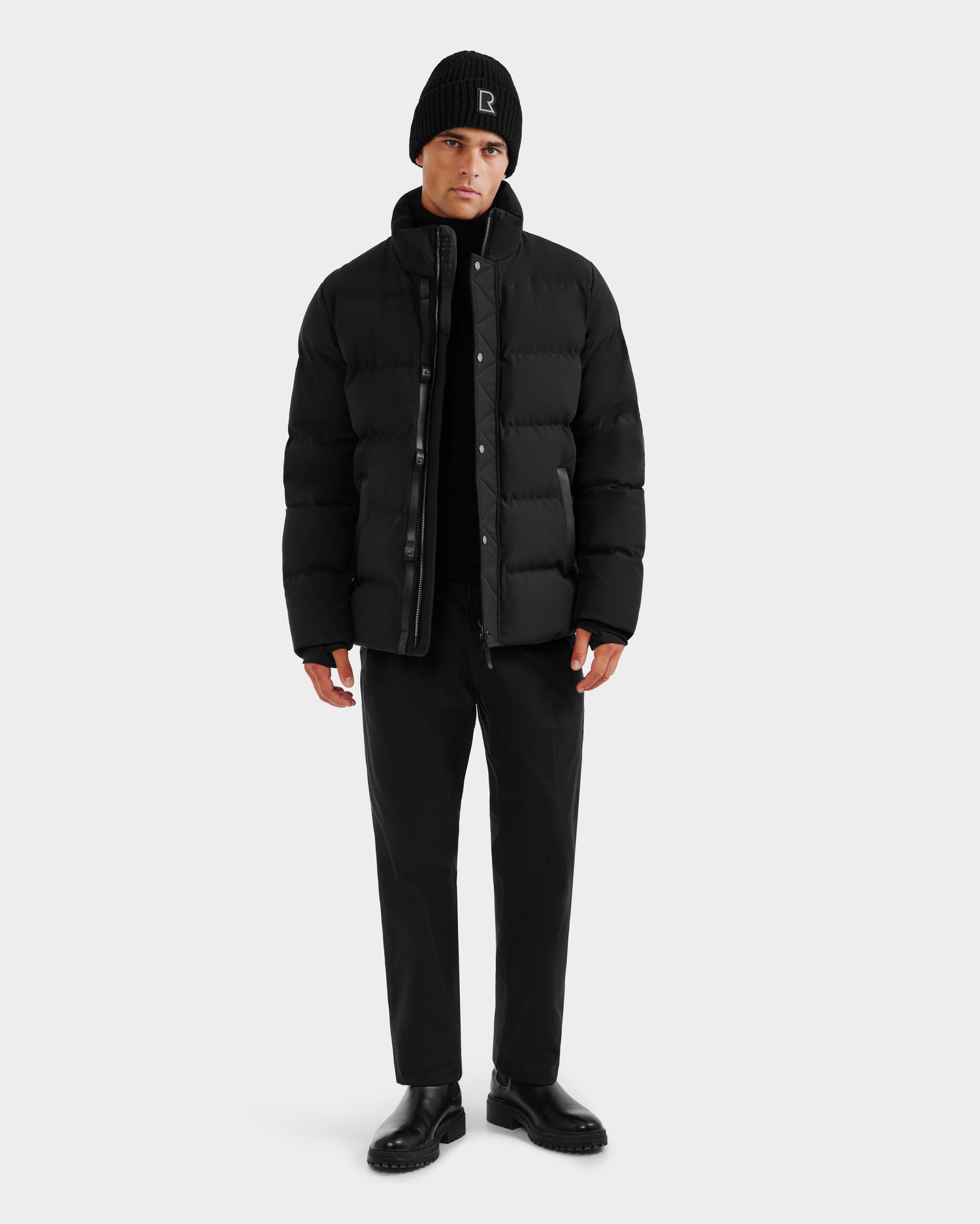 A man faces forward against a plain white background, wearing the Rudsak DOMINIC - 6125041 Black winter jacket crafted with a recycled shell, paired with black pants, shirt, and boots.