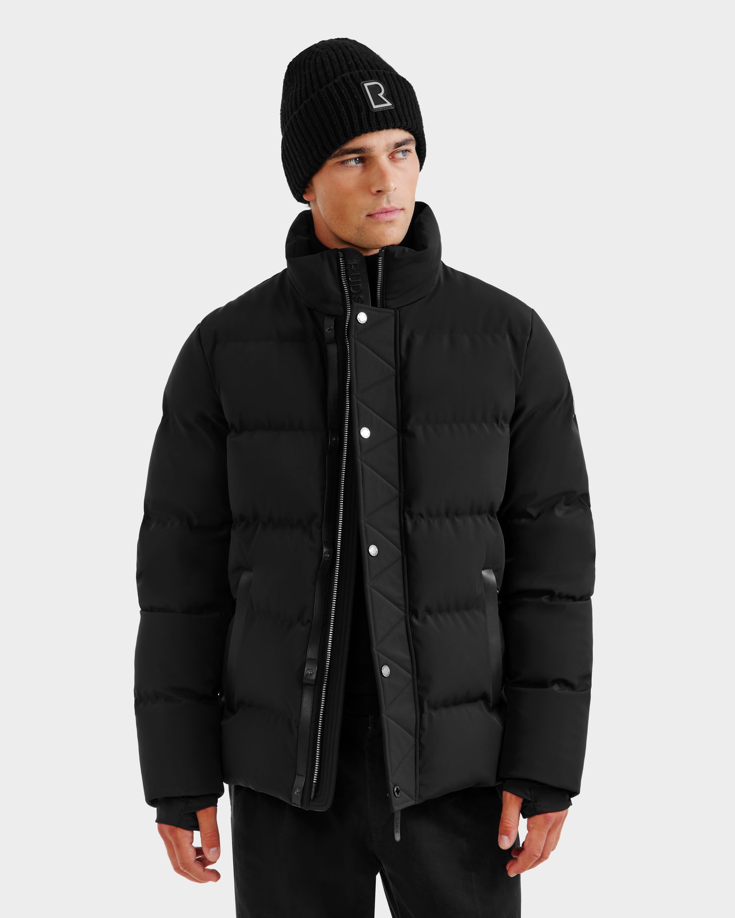 A man wearing the Rudsak DOMINIC - 6125041 Black winter jacket and black pants, paired with a black knit beanie with a white logo, stands against a light gray background looking to the side with a neutral expression.