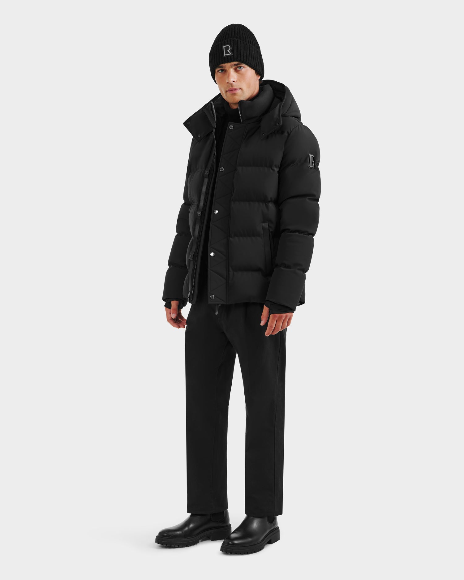 A man stands slightly left, wearing Rudsak's DOMINIC - 6125041 Black recycled shell winter jacket, a black beanie, black pants, and black boots against a plain white background.