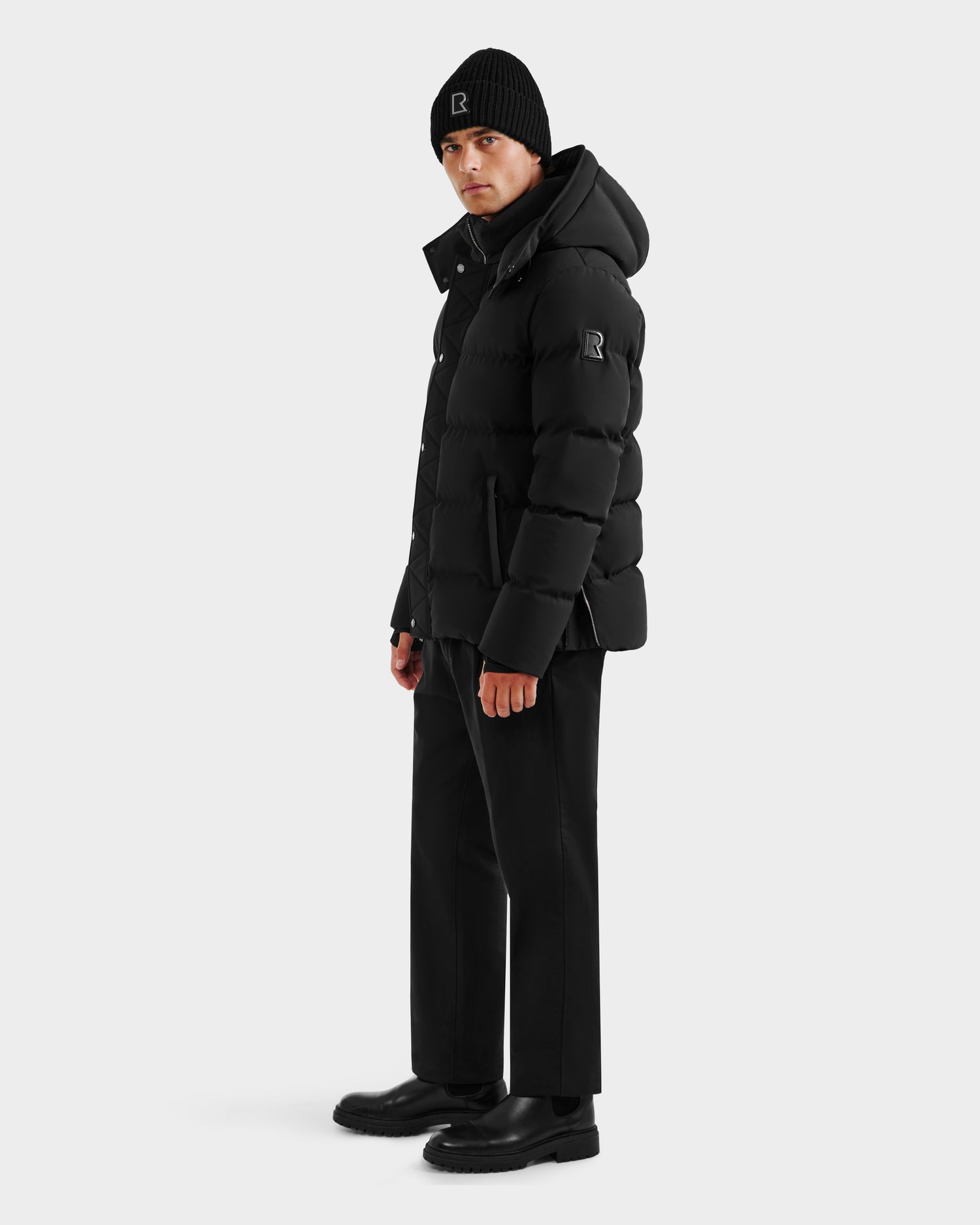 A person faces slightly left, wearing the Rudsak DOMINIC - 6125041 Black men’s down parka with a recycled shell, paired with a black beanie, black pants, and black boots, against a plain light gray background.