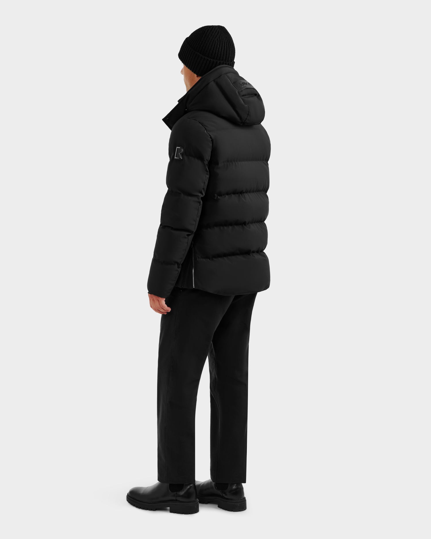 A person wearing the Rudsak DOMINIC - 6125041 Black men’s down parka stands facing away, paired with a black beanie, black pants, and black shoes against a plain light background.