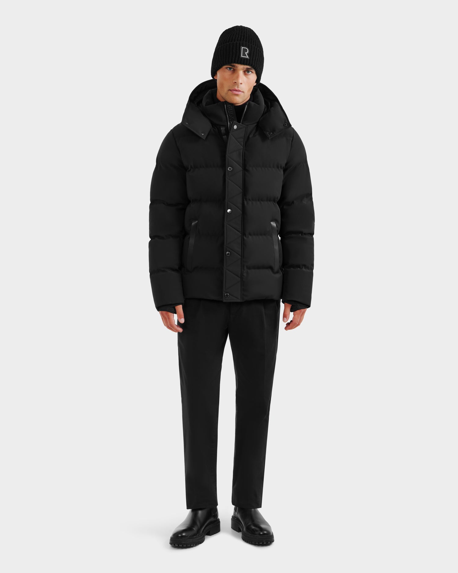 A person stands facing forward, wearing the Rudsak DOMINIC - 6125041 Black men’s down parka with a recycled shell, along with a black beanie, black pants, and black shoes against a plain white background.