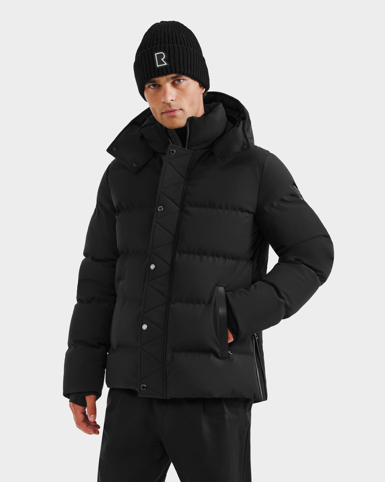A person stands against a plain light background with one hand in their pocket, wearing the Rudsak DOMINIC - 6125041 Black men’s down parka and black pants, paired with a black knit beanie featuring a white R on the front.