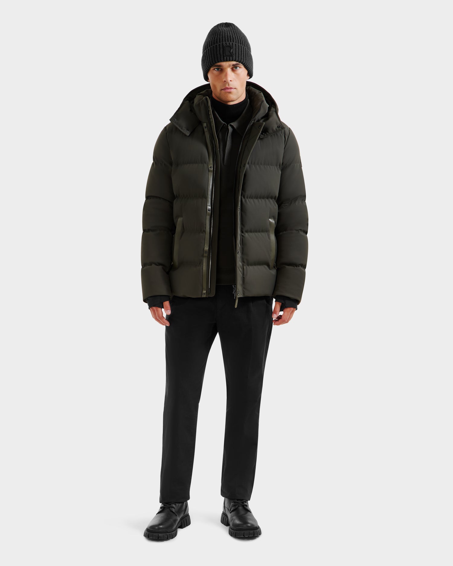 A person faces forward, wearing the Rudsak DOMINIC - 6125041 Dark Olive men’s down parka with a recycled shell, a black beanie, black pants, and shoes against a plain light background.