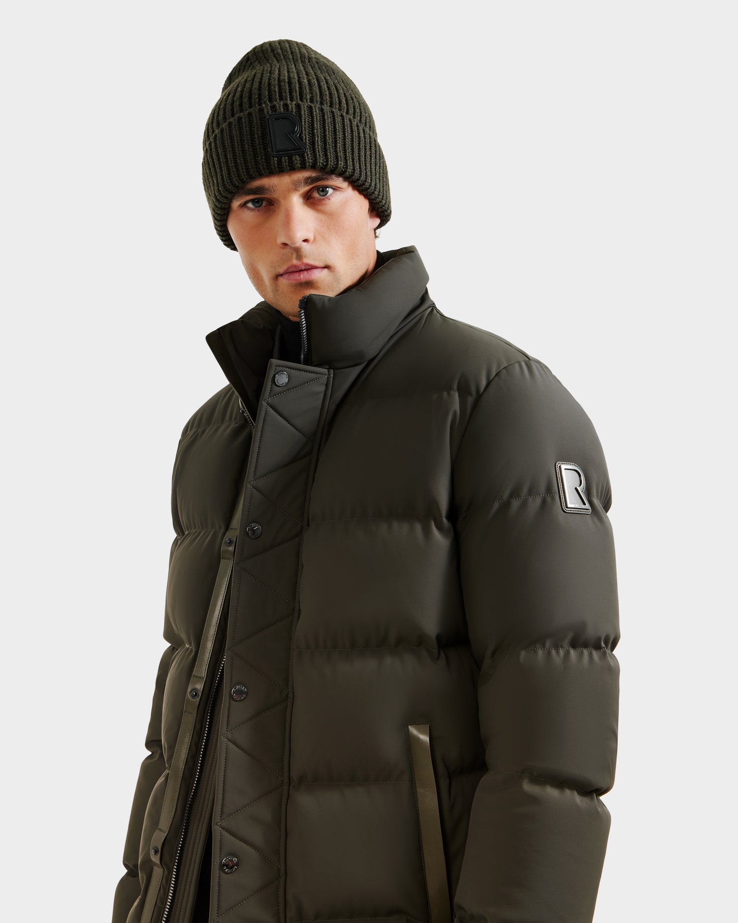 A man wearing the Rudsak DOMINIC - 6125041 Dark Olive men’s down parka and a matching knit beanie looks into the camera against a plain white background.