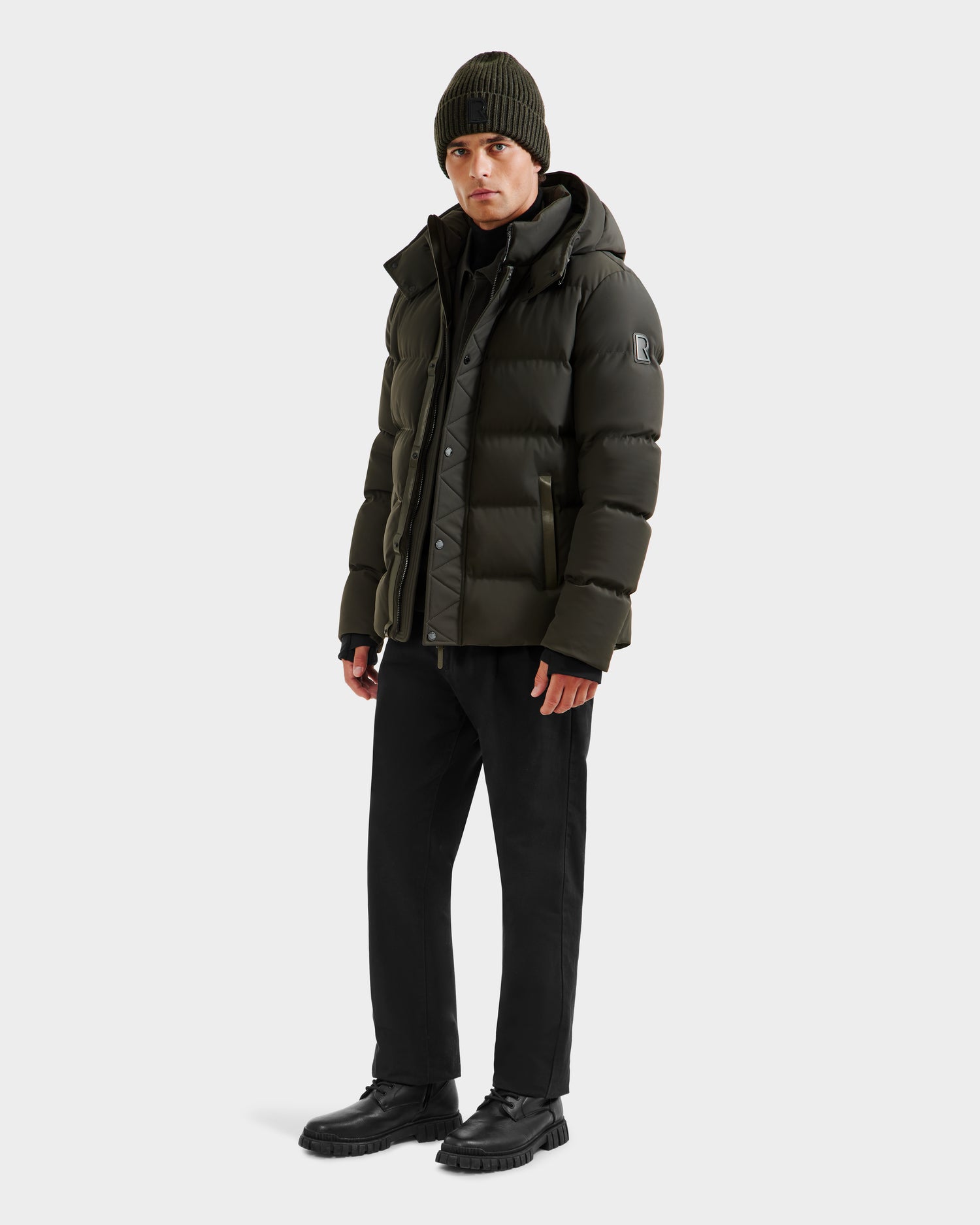 A person wears the Rudsak DOMINIC - 6125041 Dark Olive men’s down parka, featuring a recycled shell, paired with black pants, black shoes, and a green knit beanie, standing against a plain white background.