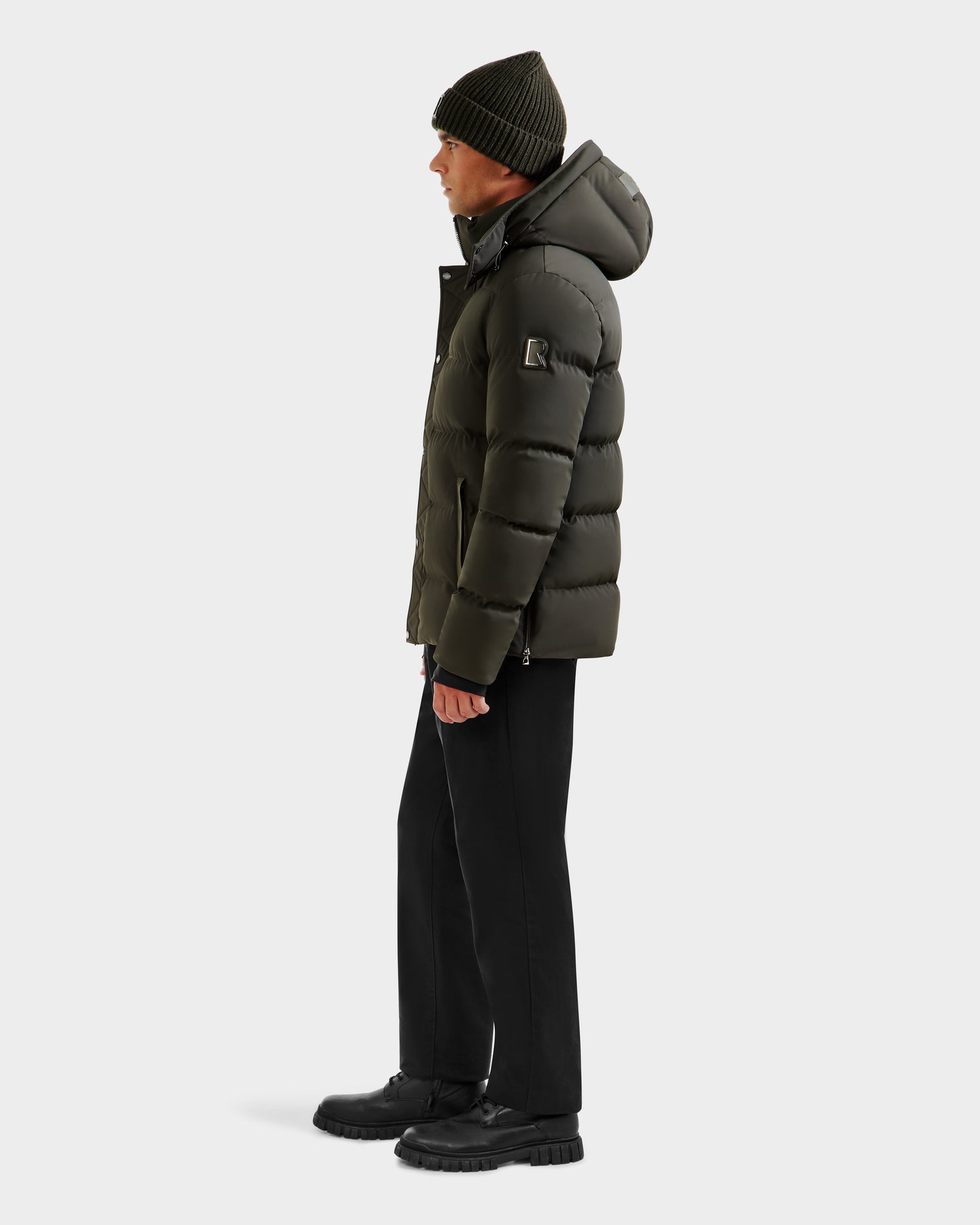 A person in profile wears the Rudsak DOMINIC - 6125041 Dark Olive men’s down parka with recycled shell, black pants and shoes, plus a dark knit beanie, set against a plain white background.