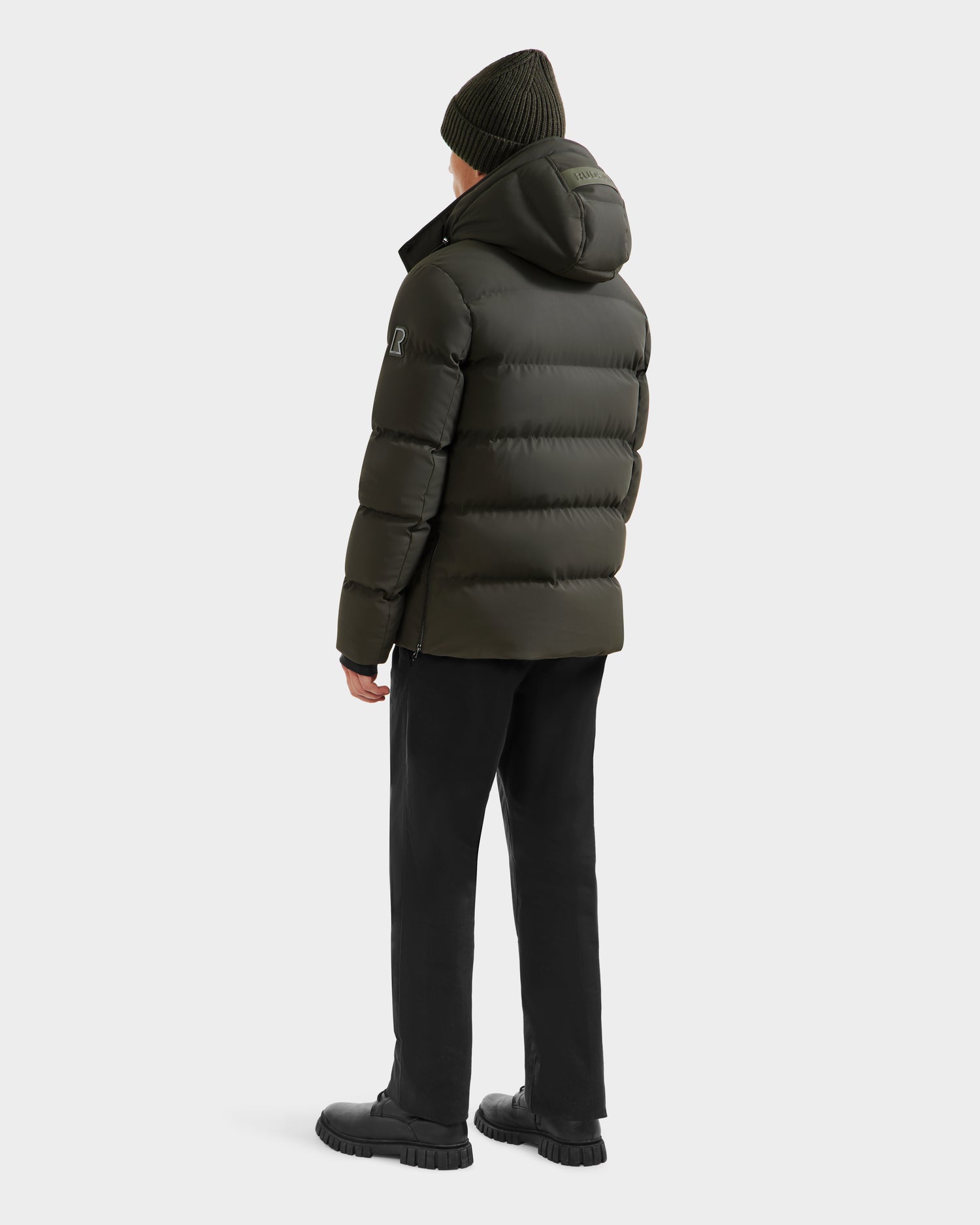 A person in a Rudsak DOMINIC - 6125041 Dark Olive men’s down parka with recycled shell and matching beanie, paired with black pants and boots, stands facing away against a plain light background.