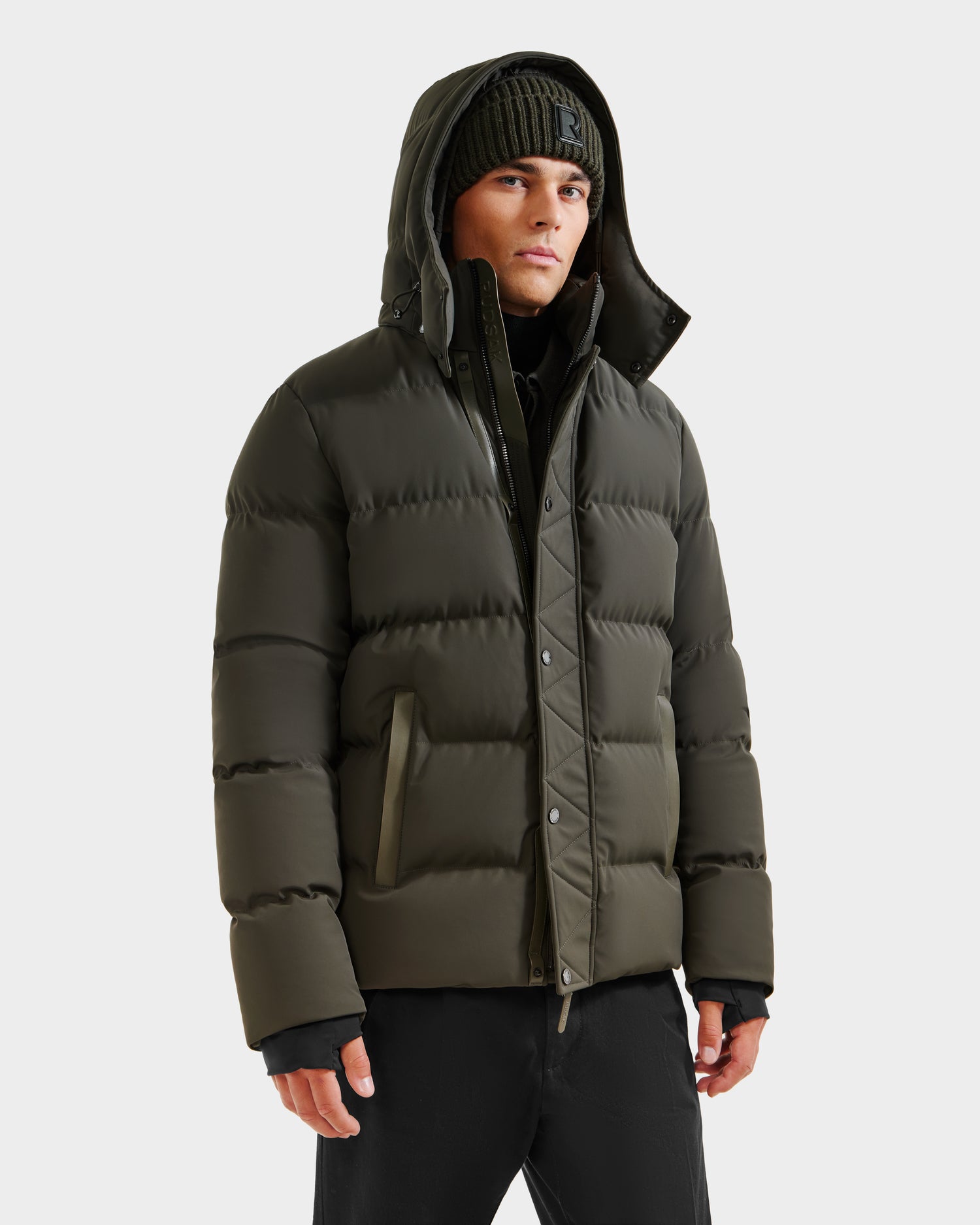 A person wearing the Rudsak DOMINIC - 6125041 Dark Olive weather-resistant winter jacket with a hood, black pants, and a green knit beanie, stands against a plain light background.