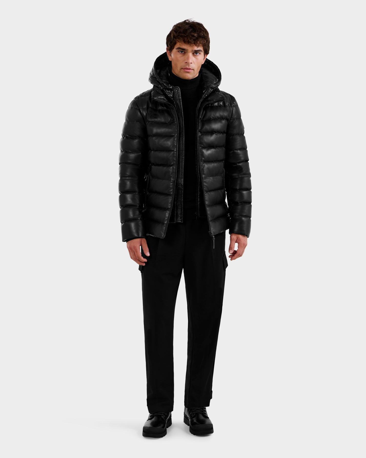 A man stands facing forward against a white background, wearing the Rudsak DRAVENX - 6125686 Black men's leather down jacket with a hood, paired with a black turtleneck, pants, and shoes.