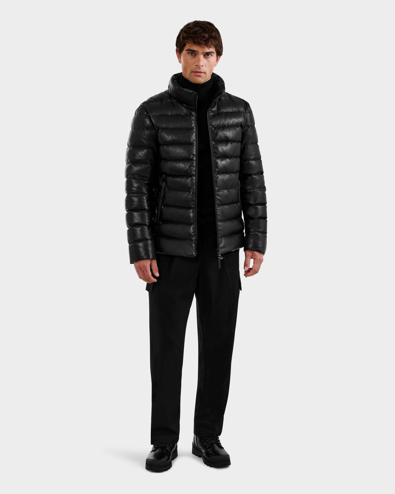A man stands against a plain white background wearing a sleek black Rudsak DRAVENX - 6125686 leather puffer jacket, paired with a black turtleneck, pants, and shoes. He faces forward with a neutral expression.