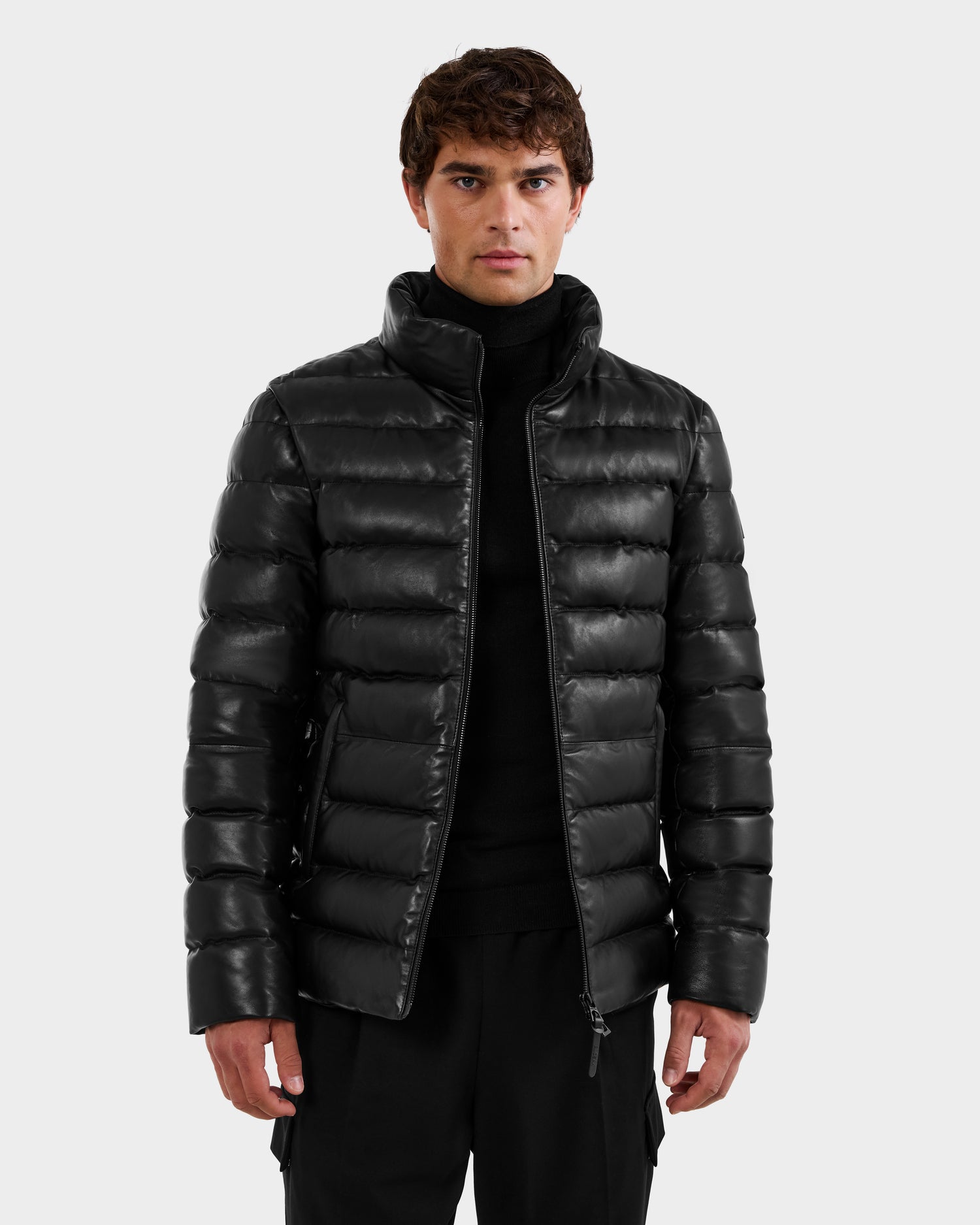 A man wearing the Rudsak DRAVENX - 6125686 Black leather puffer over a black turtleneck stands facing forward against a plain light gray background.