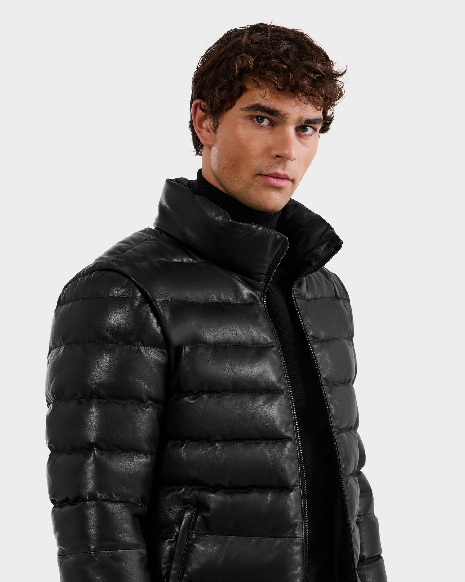 A young man with curly brown hair wears the Rudsak DRAVENX - 6125686 Black leather puffer jacket over a black turtleneck, standing against a plain light gray background and looking at the camera with a neutral expression.