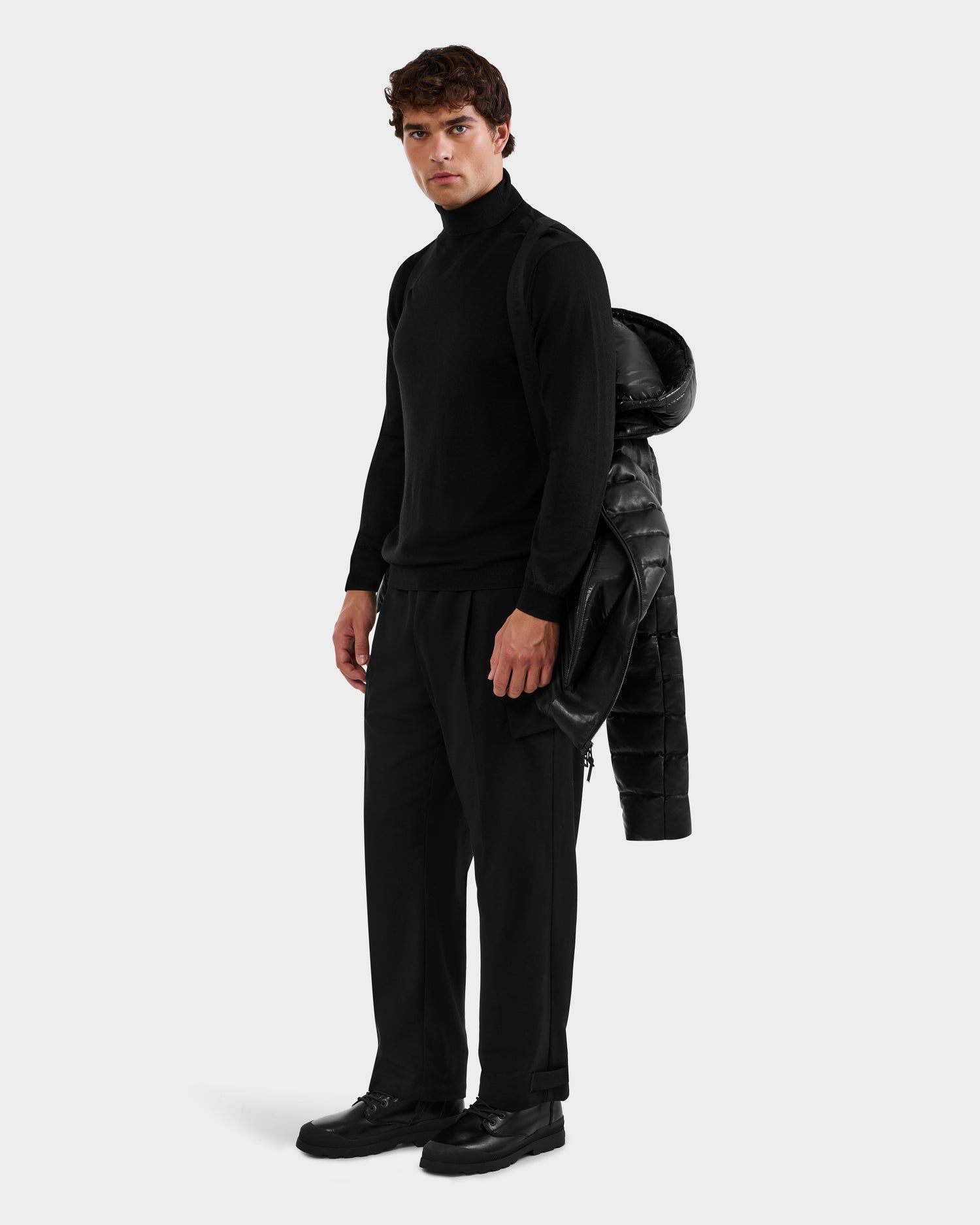 A man stands against a plain background wearing black attire and holding a Rudsak DRAVENX - 6125686 Black men’s leather down jacket draped over his left arm.